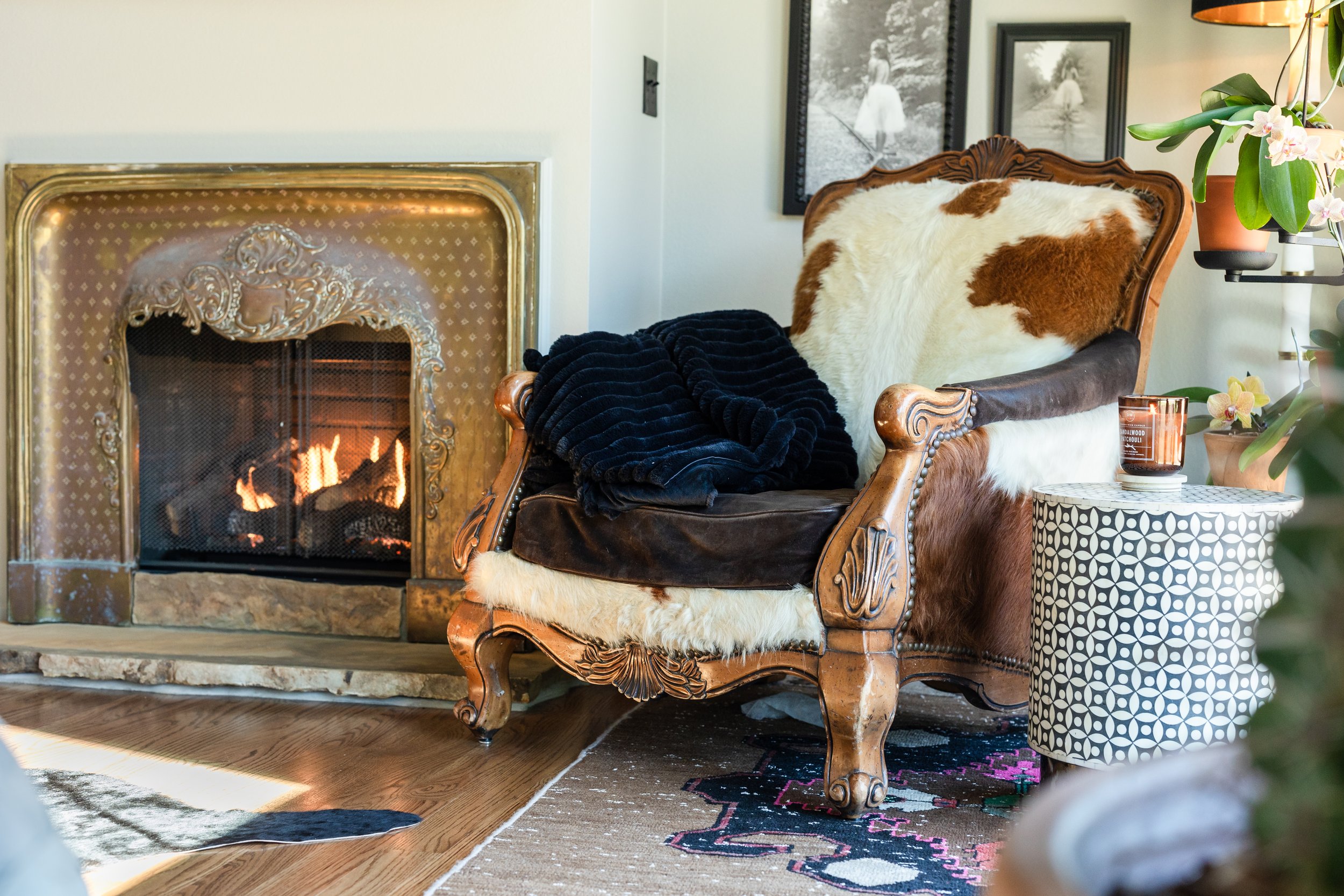 A cozy living room corner with a vintage wooden armchair covered with a cowhide blanket and a black plush throw, next to a fireplace with carved gold trim and a fire burning inside. There are framed black and white photographs on the wall, a potted o
