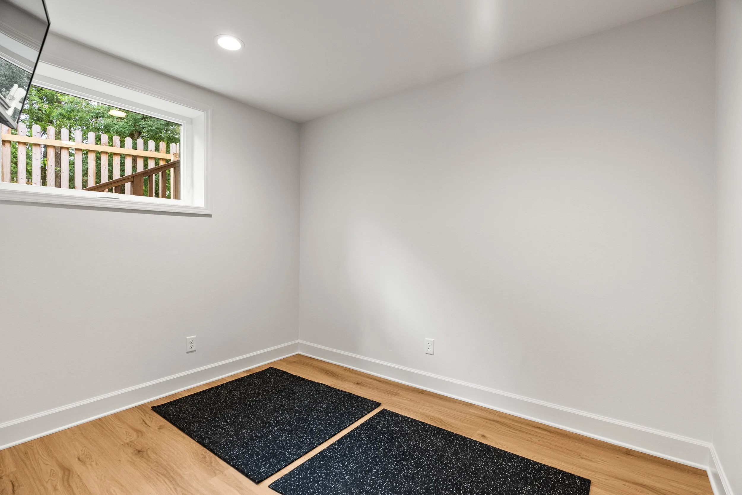 Empty room with white walls, hardwood floor, small window, and black rugs near the wall.