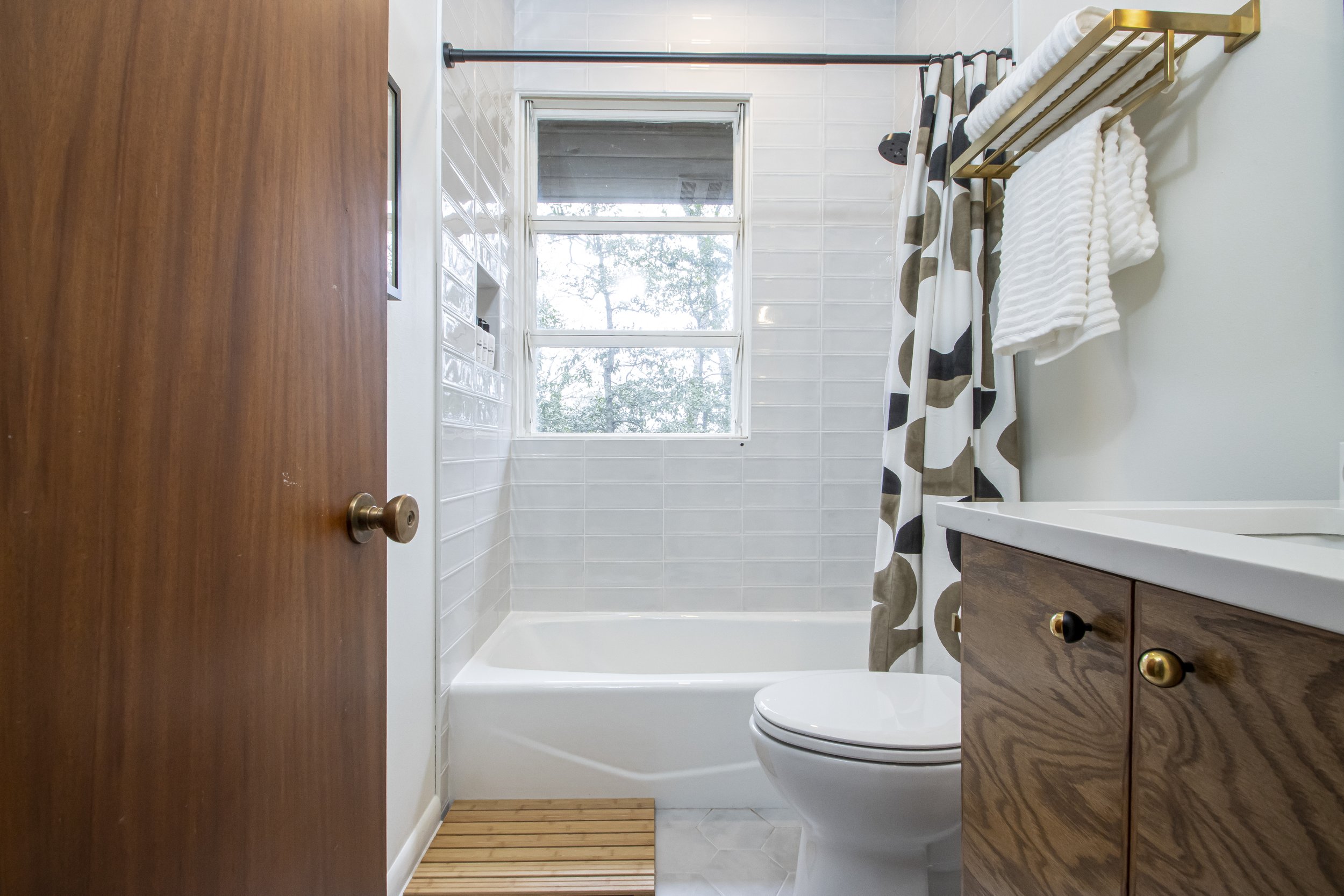 View of a small bathroom with a bathtub and a window with trees outside. The bathroom has white tiled walls, wooden cabinetry, and a striped shower curtain. There are towels on a golden towel rack and a wooden bath mat in front of the tub.