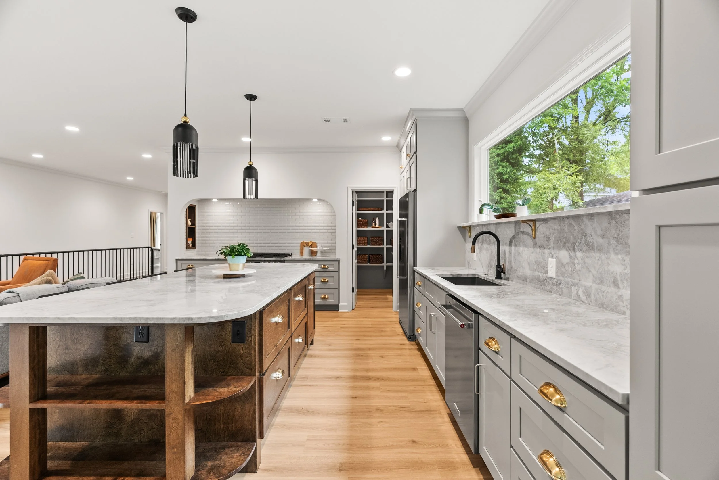 Modern kitchen with white cabinets, marble countertops, black pendant lights, large window showing greenery outside, and hardwood floors.