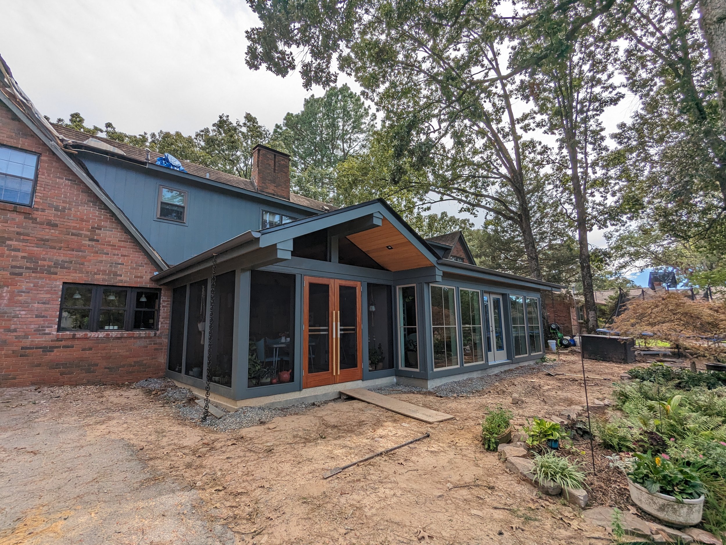 The image shows the back of a brick house with a new screened-in porch attached. The house has a sloped roof with a chimney, and there are large trees surrounding the property. The yard appears to be under landscaping or construction, with dirt and s