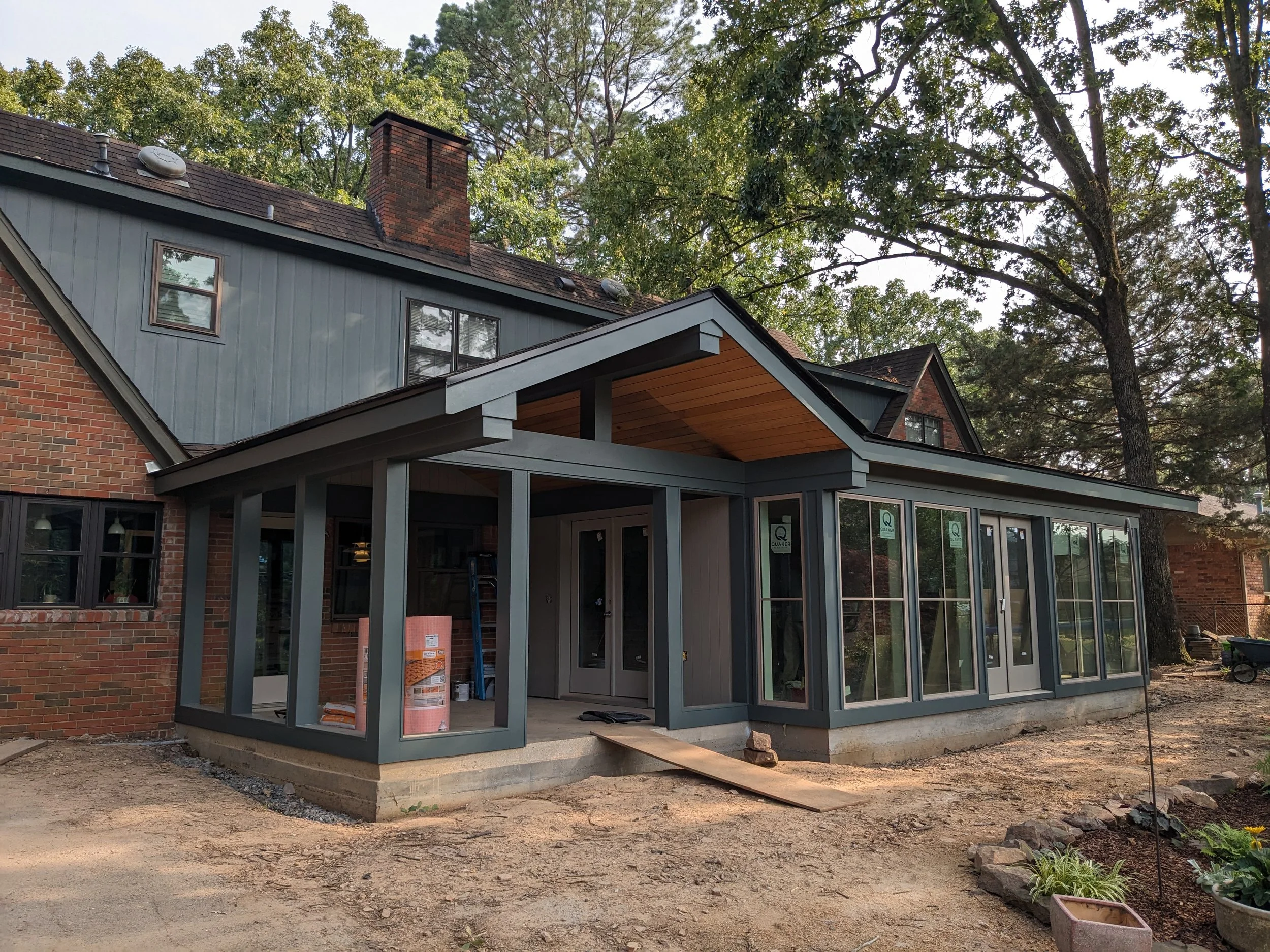 A house under construction featuring a brick section and a modern glass extension with a patio. The house has a brick chimney and a gray exterior. There are trees with green leaves surrounding the house.