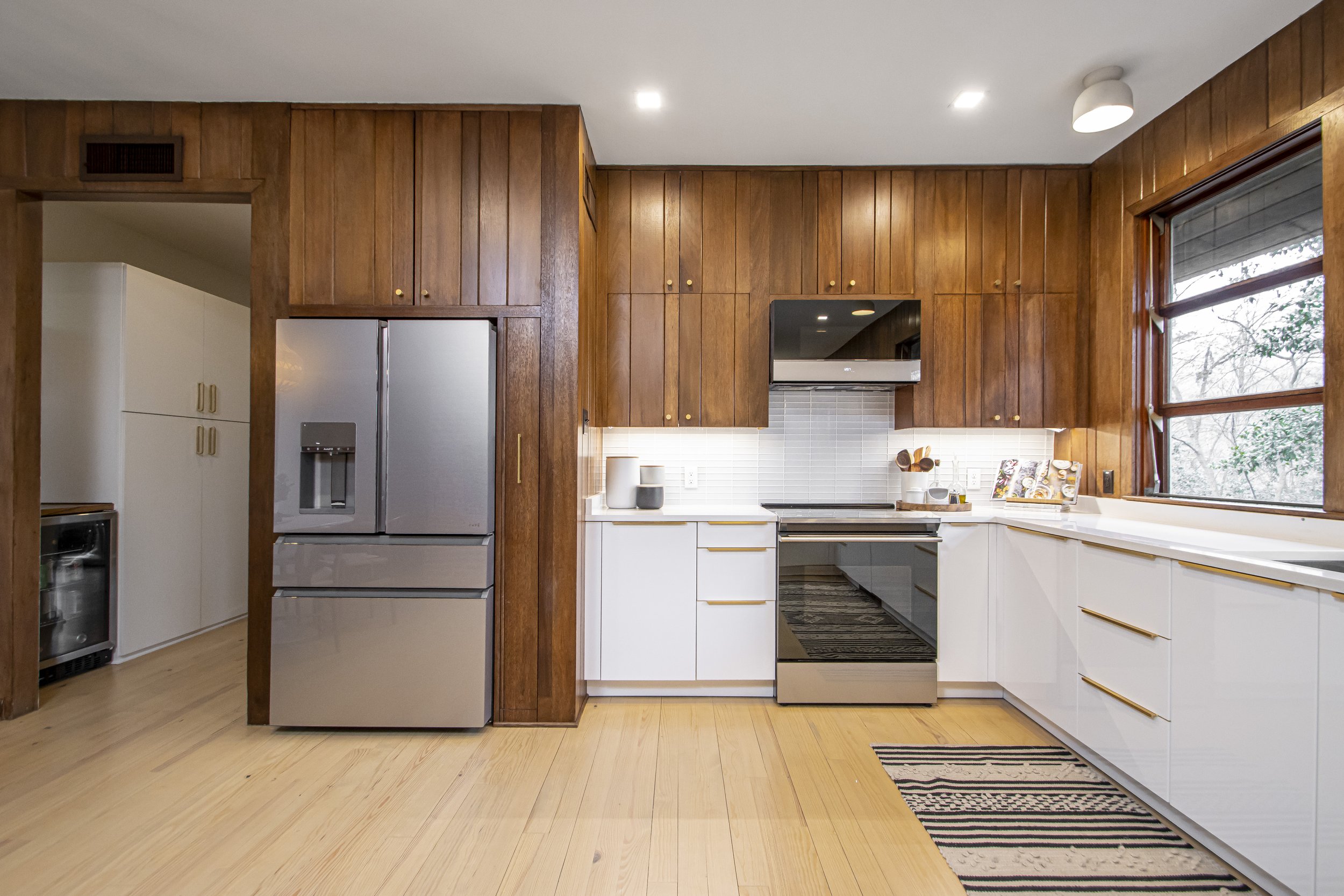 Kitchen with wood-paneled upper cabinets, white lower cabinets, stainless steel refrigerator, black stove, and a window showing outdoor trees.