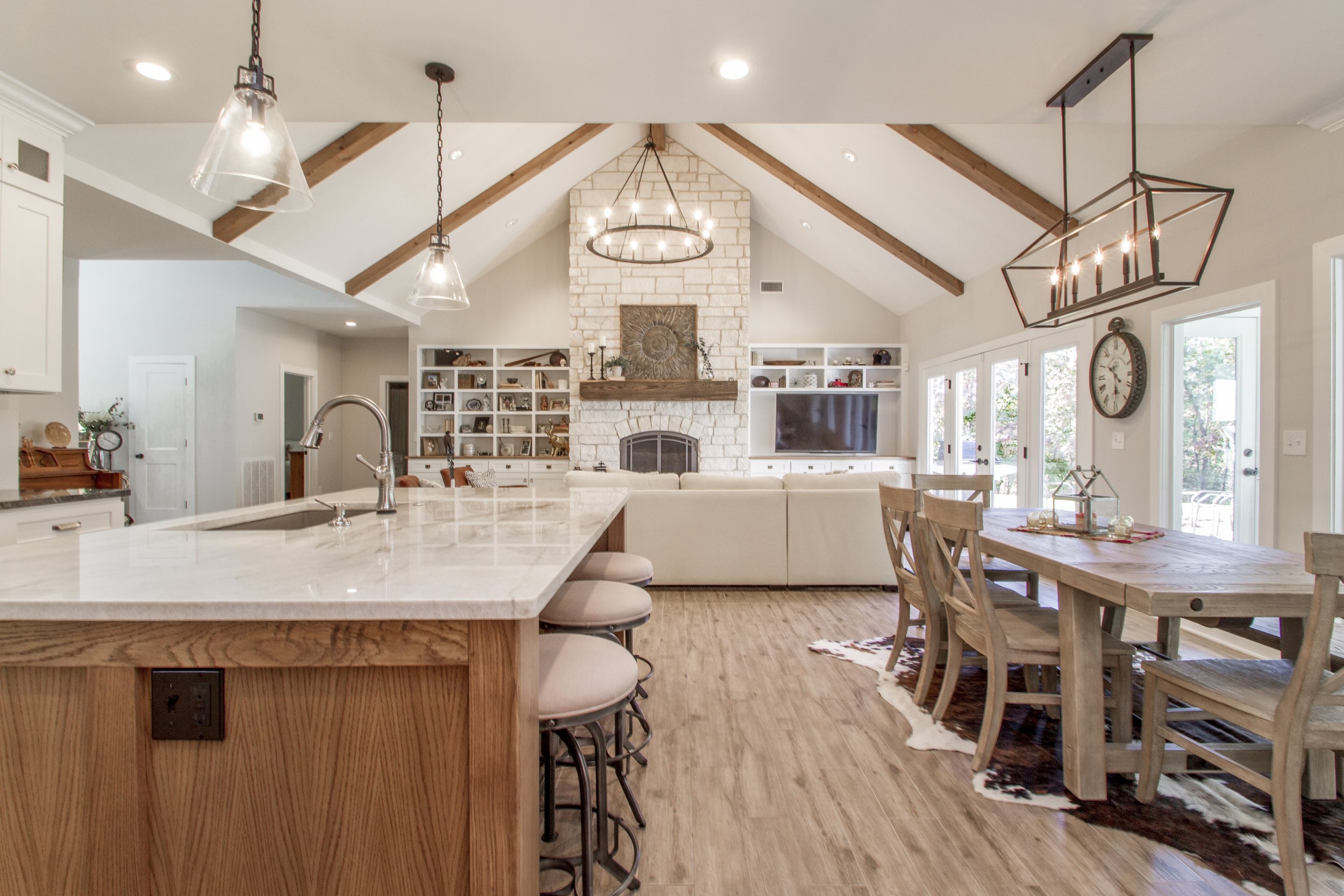Open floor plan kitchen and living room with vaulted ceilings, wooden beams, and a stone fireplace, featuring a kitchen island with bar stools, a dining table, and a large wall-mounted TV.