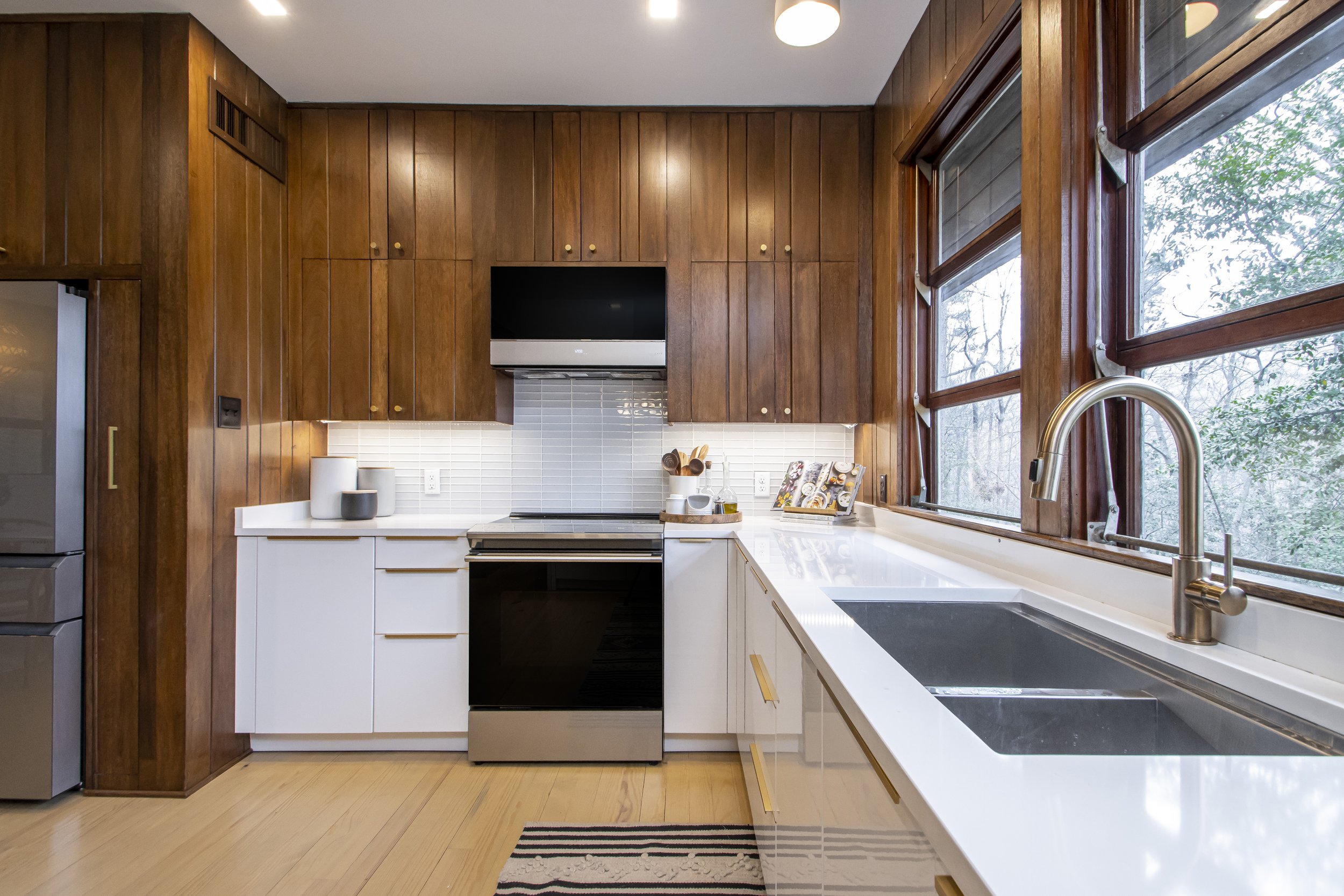 Kitchen with white countertops, wooden cabinets, and large windows showing trees outside. Includes a faucet and some small kitchen items on the counter.