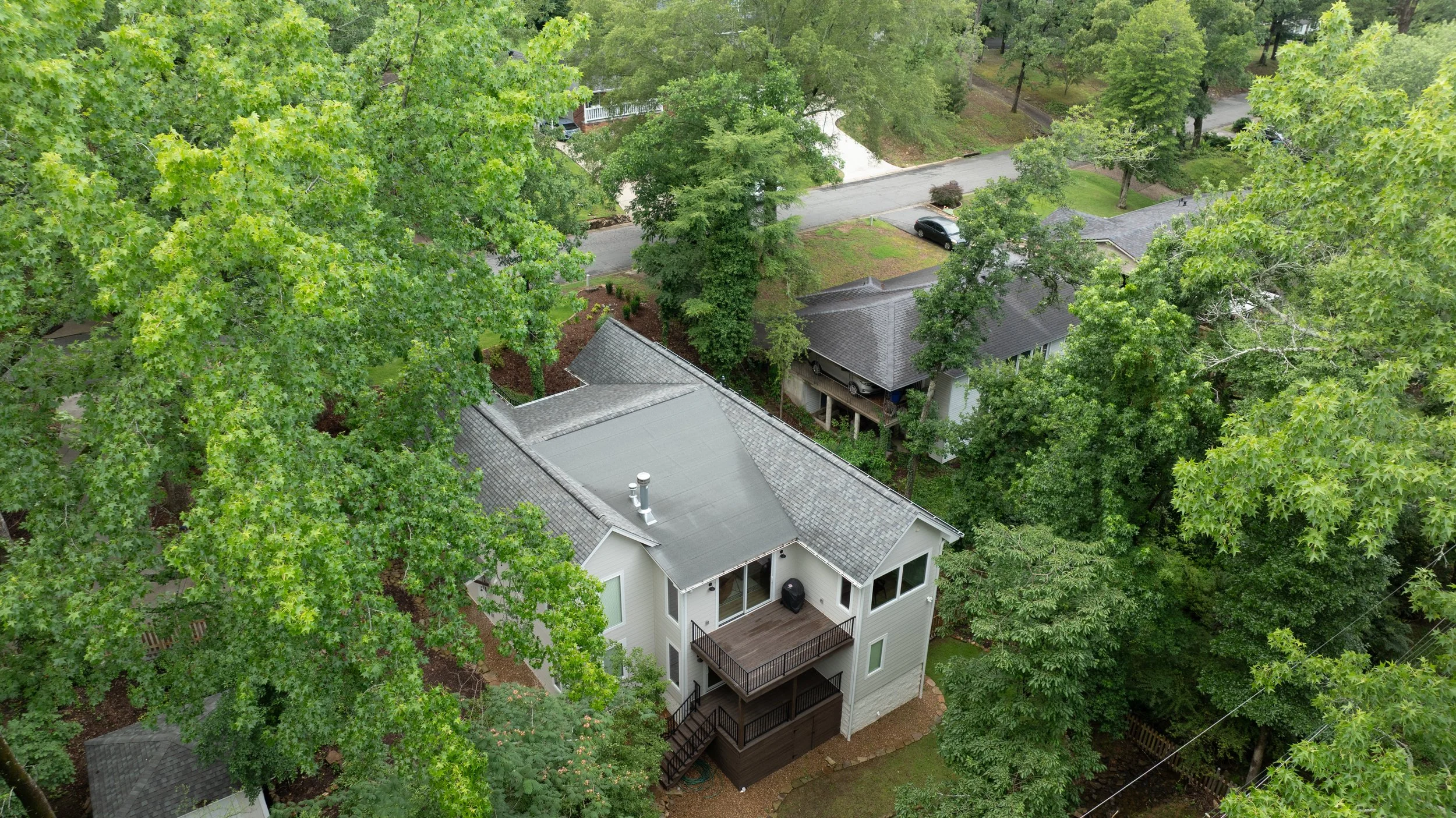 Aerial view of a residential neighborhood with large trees, houses, and a street with parked cars.