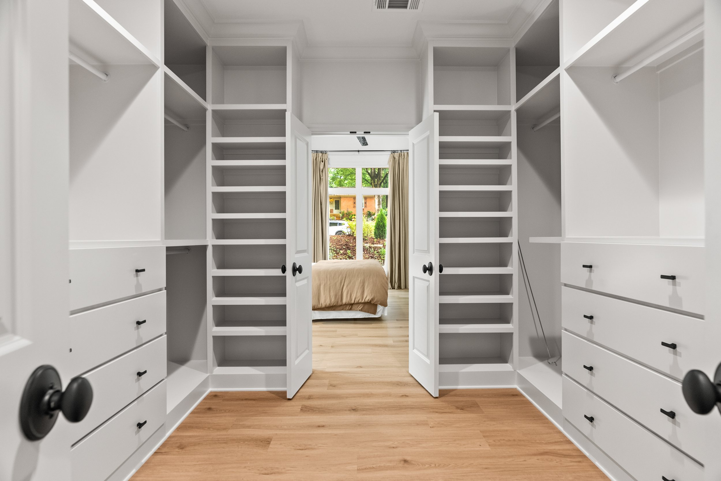 A walk-in closet with white built-in shelves and drawers, and a doorway leading to a bedroom with a window and beige curtains, view of a tree and houses outside.