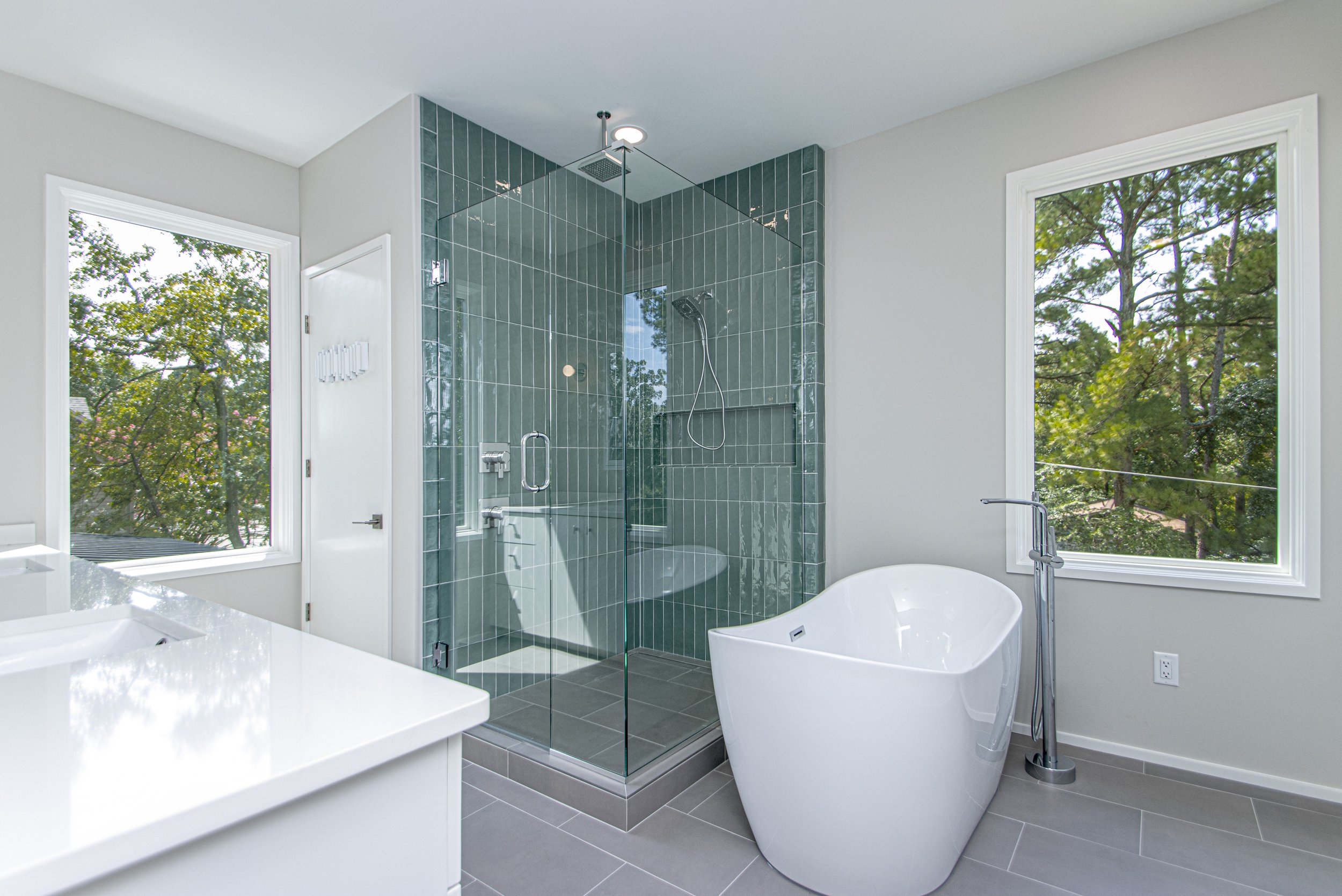 A modern bathroom featuring a glass-enclosed shower with green tiles, a freestanding bathtub, two large windows showing trees outside, and white walls with minimal decor.