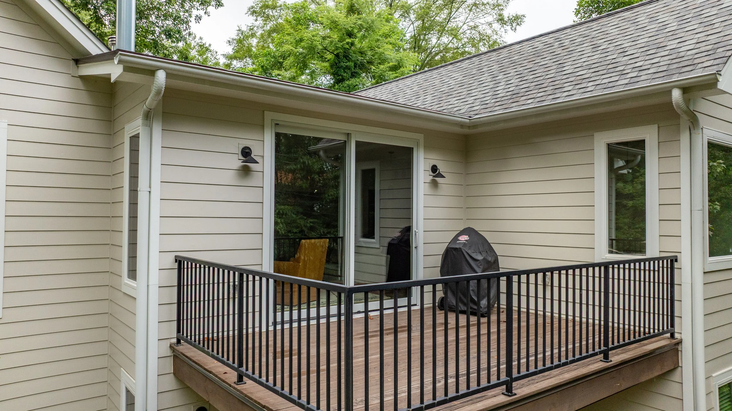A small balcony with a black metal railing attached to a beige house. There is a grill covered with a black protective cover and a yellow chair on the balcony. The house has beige siding and two black outdoor wall lights. The background shows green t