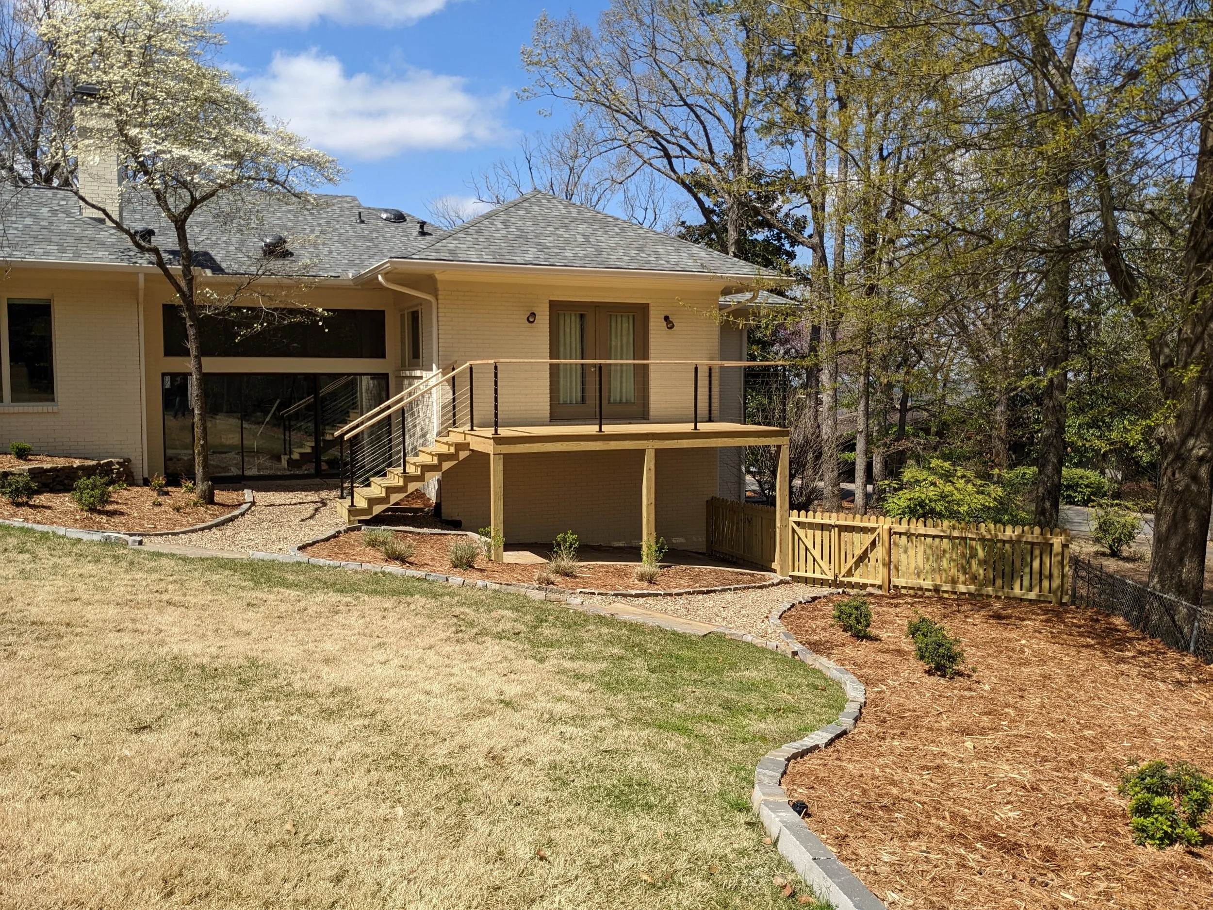 A backyard with a white brick house, a new wooden deck with stairs and black railing, trees with some blossoms and green leaves, a grassy lawn, and a stone pathway edged with mulch.