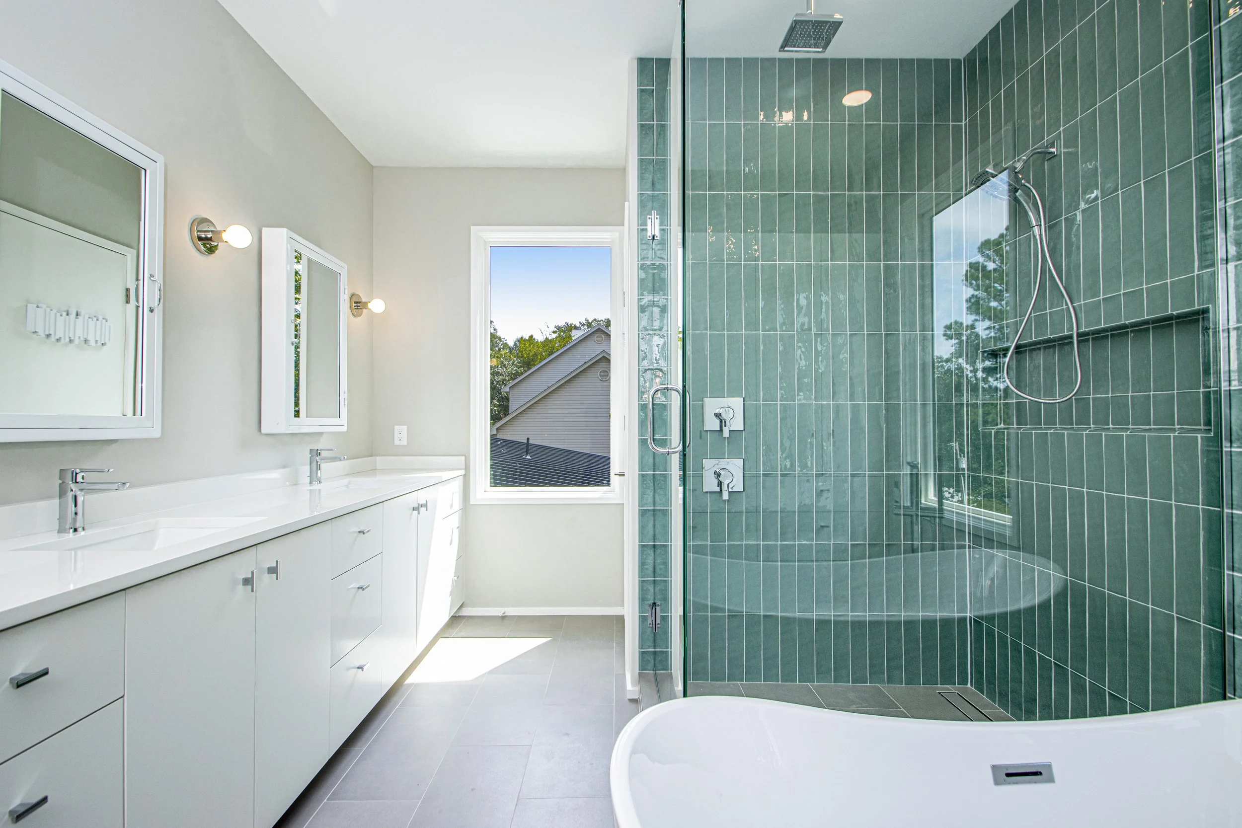Modern bathroom with white vanity, dual sinks, large mirror, wall-mounted light fixtures, and a walk-in shower with green tiles and a glass door, illuminated by natural light from a window.
