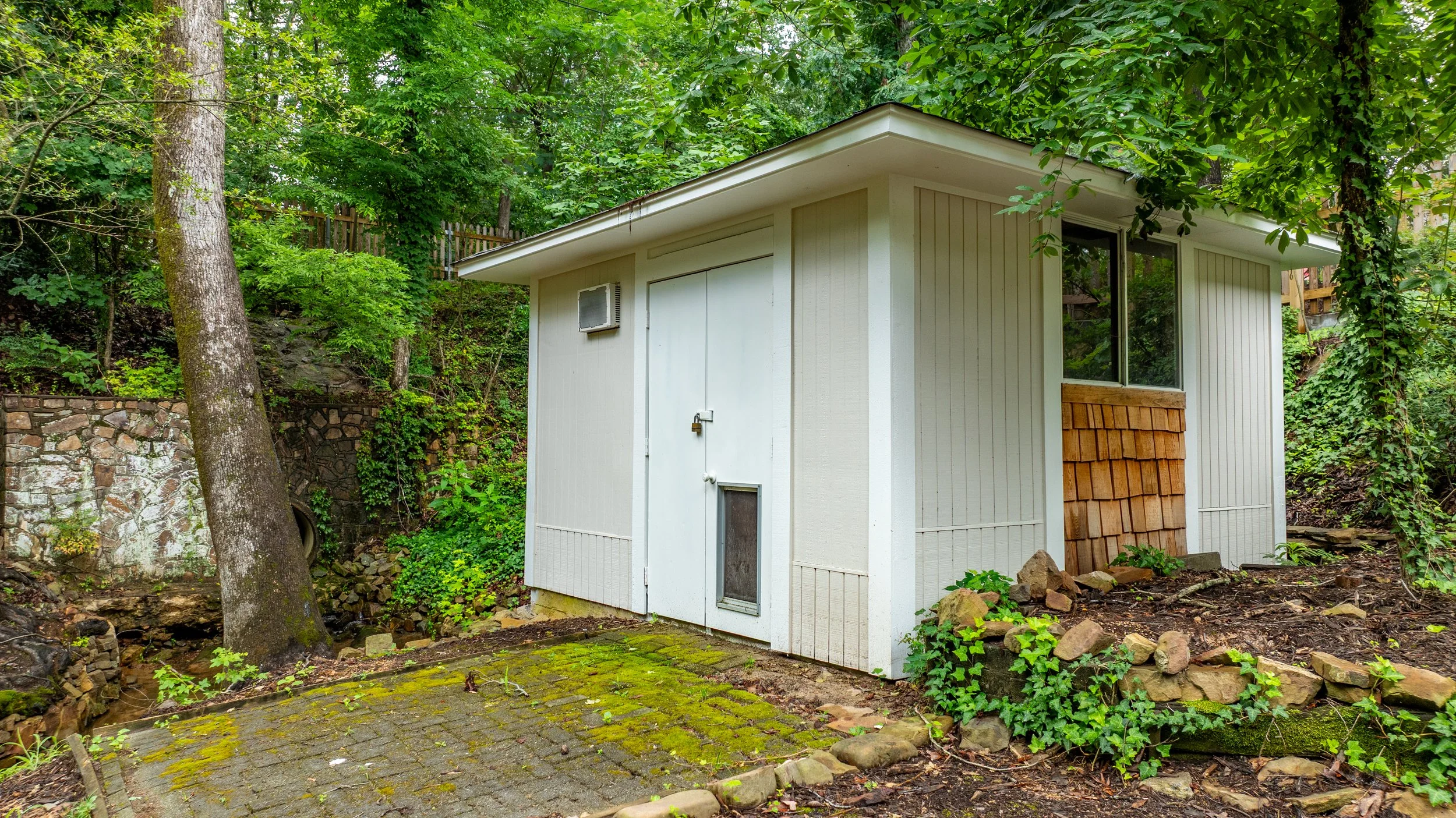 A small white shed with a window and a vent, surrounded by green trees and plants, with a stone and moss-covered paved area in the foreground.