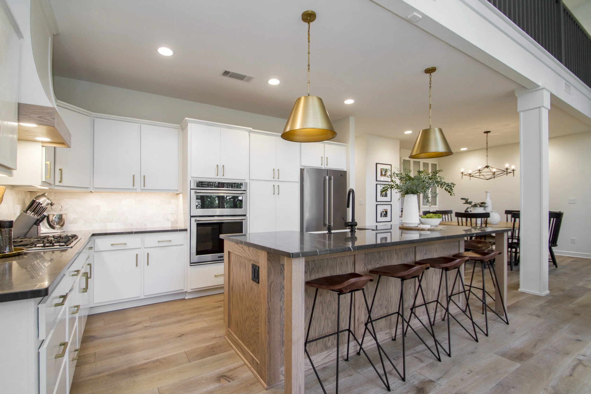 Modern kitchen with white cabinets, stainless steel appliances, and a large wooden island with black barstools, pendant lighting, and a dining area in the background.