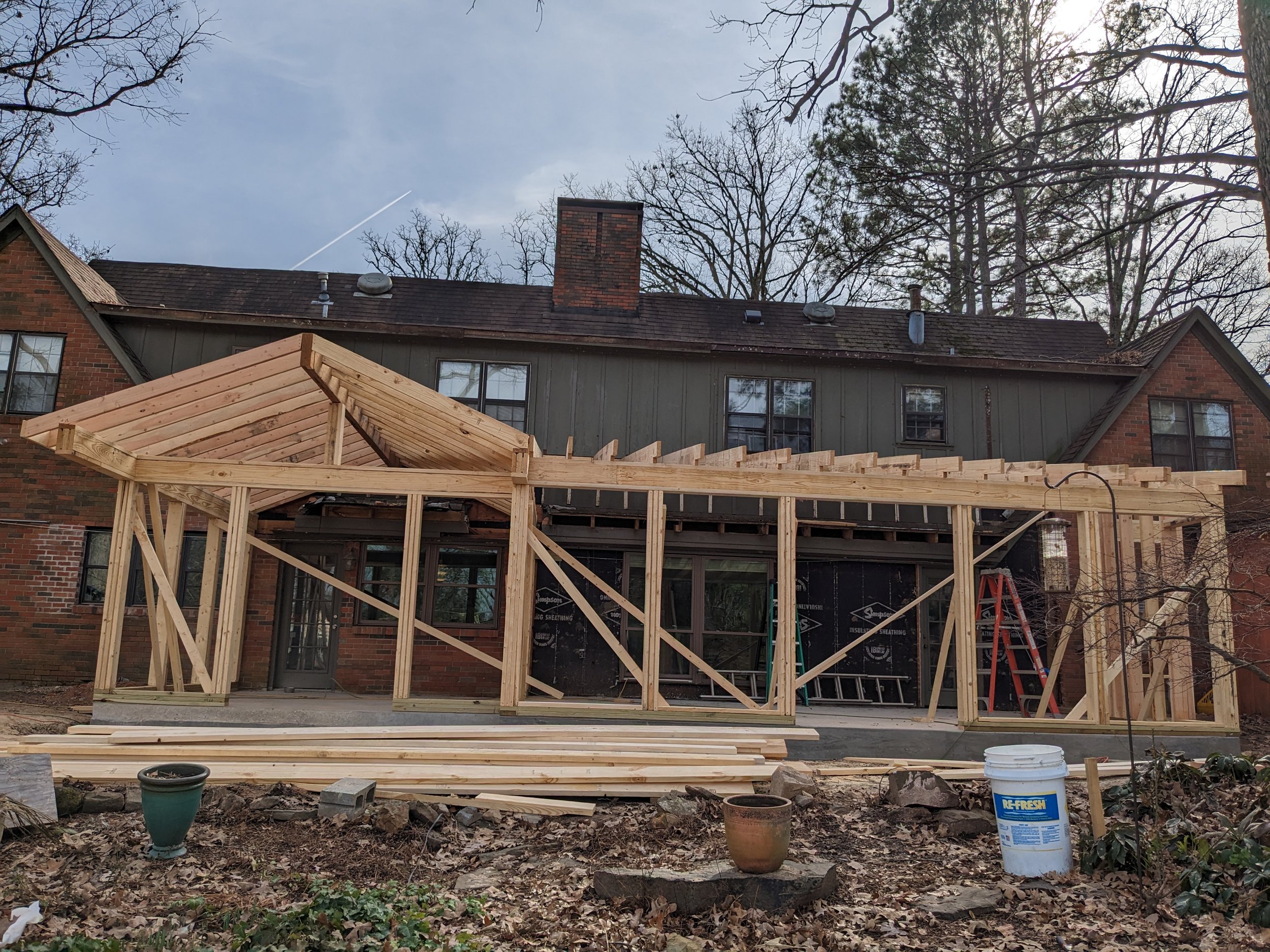 Residential backyard under construction with a wooden deck frame, potted plants, buckets, and a house with brick and gray siding. Trees are visible in the background and the sky is cloudy.