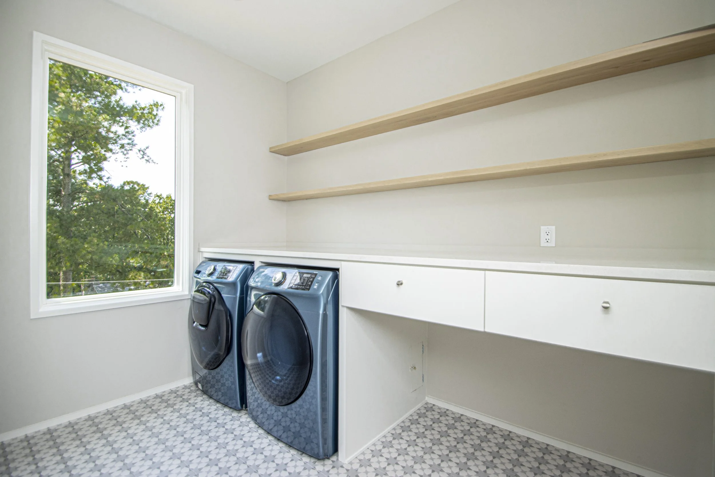 Laundry room with a large window, two blue front-loading washing machines, white cabinets, floating shelves, and patterned tile floor.