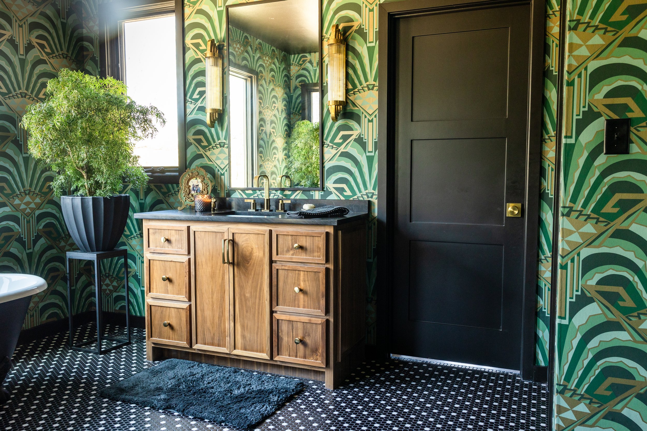 A bathroom with geometric green and gold wallpaper, a black framed mirror, a wooden vanity with a black countertop, a large potted plant, and vintage-style gold wall sconces, with black and white patterned tile floor and black door.