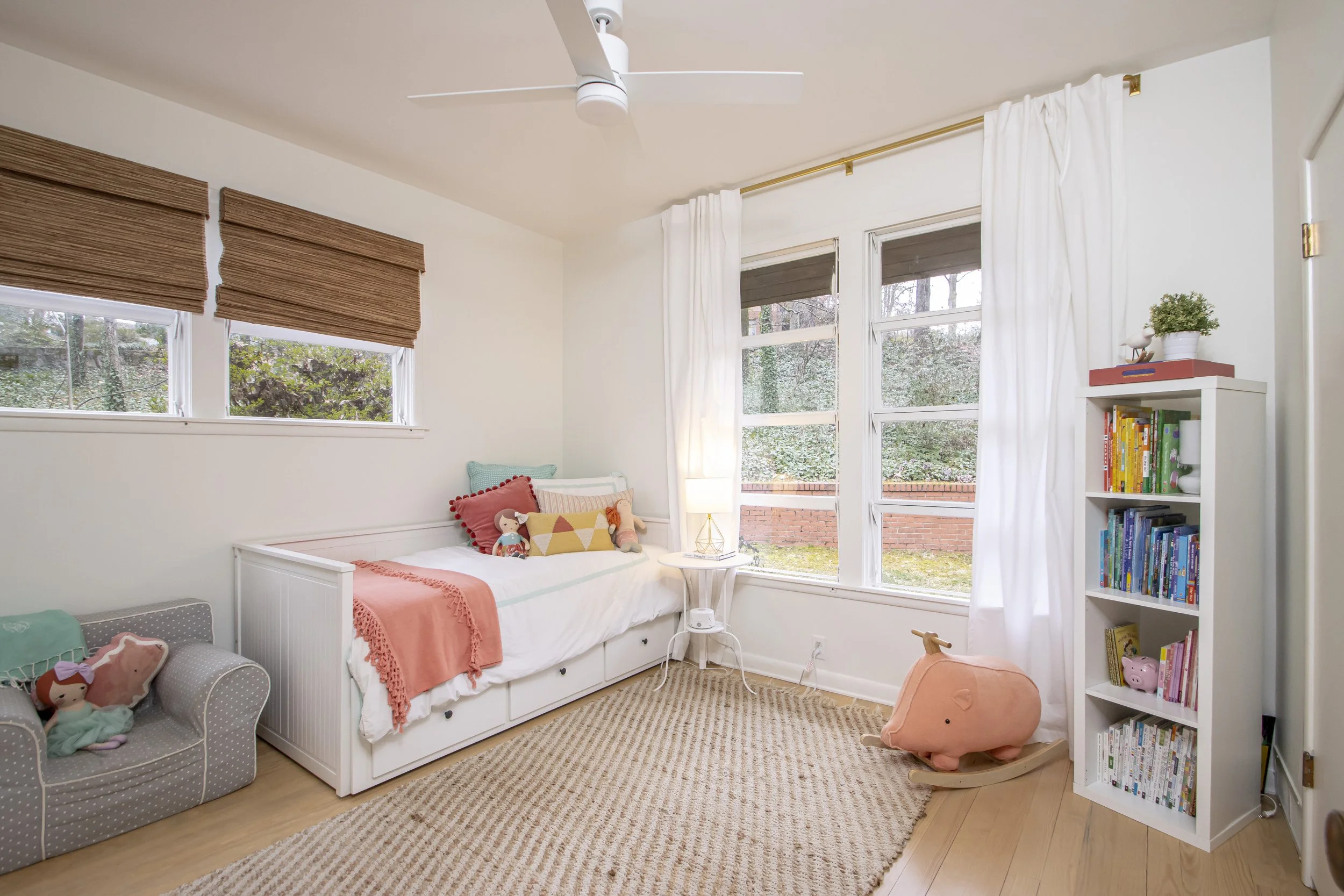 A cozy bedroom with white walls and large windows with white curtains, a white bed with colorful pillows, a beige rug, and a white bookshelf filled with books, a decorative plant, and toys. There is a fan on the ceiling.