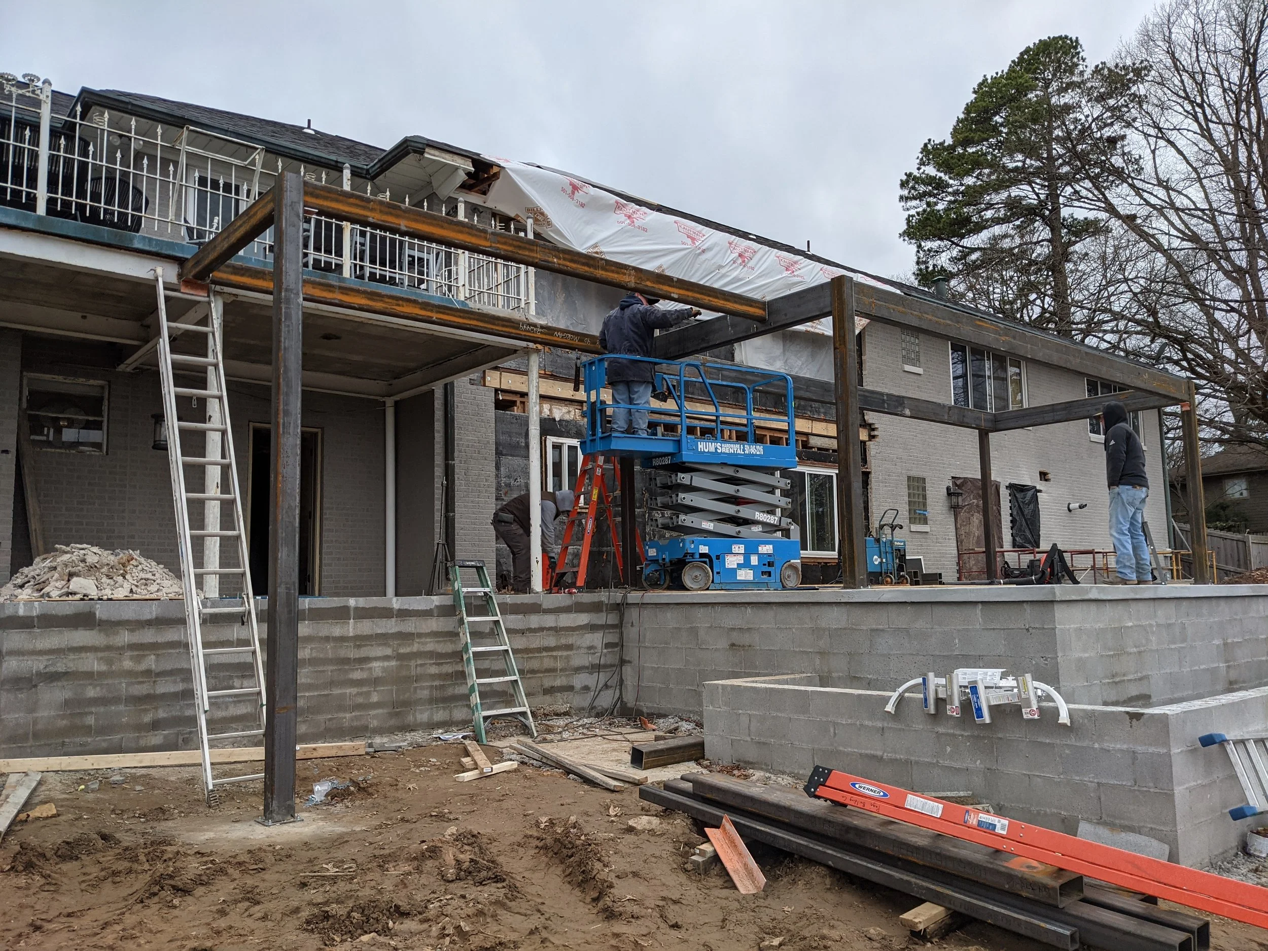 Construction workers building a deck outside a two-story house, with scaffolding and construction materials on site.