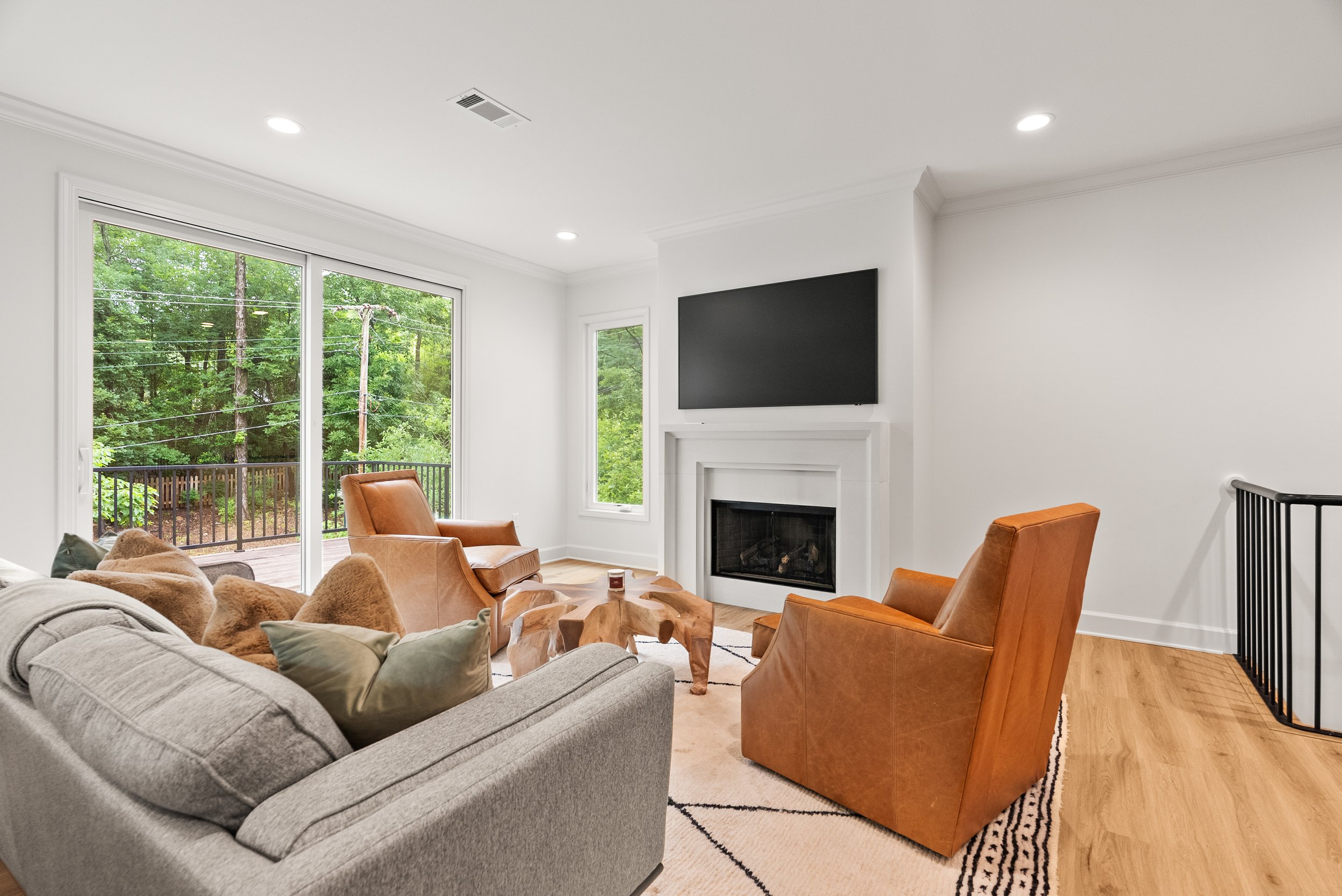 Living room with large glass sliding door showing green trees outside, white walls, a fireplace, a mounted flat-screen TV, gray sofa with pillows, tan leather armchairs, and a wooden coffee table.