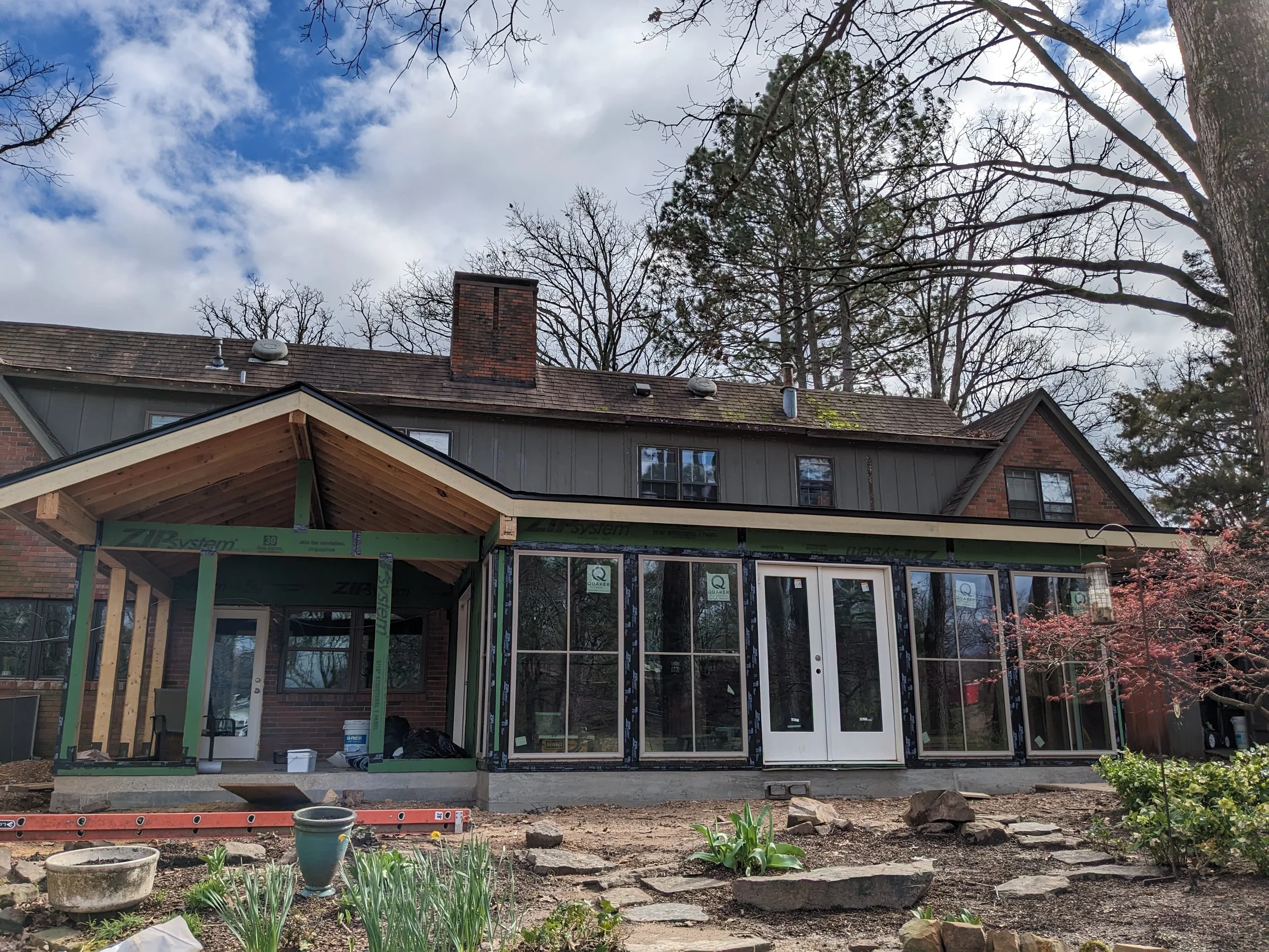 Construction of a sunroom or extension on the back of a brick house, with a partially built structure featuring a gable roof, front windows, and an unfinished frame on the left side, set in a backyard with trees and a cloudy sky.