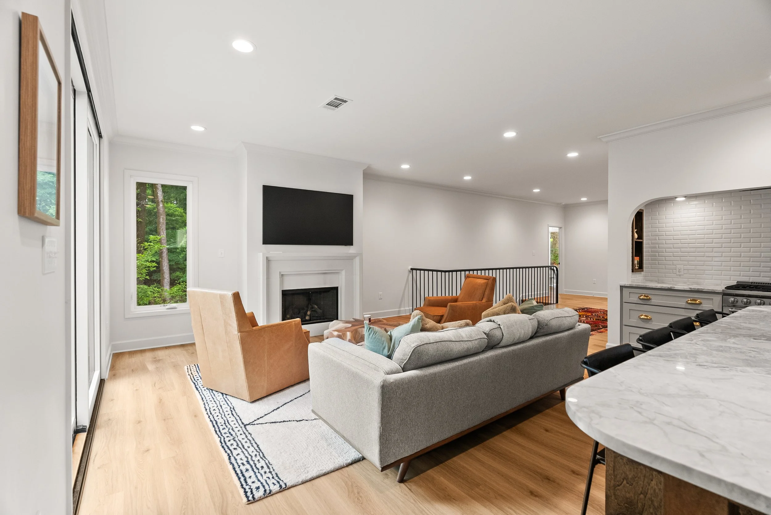 Living room with beige sofa and plaid armchair, mounted flat-screen TV above fireplace, white walls, hardwood flooring, large window with greenery outside, part of kitchen with marble countertop and bar stools.