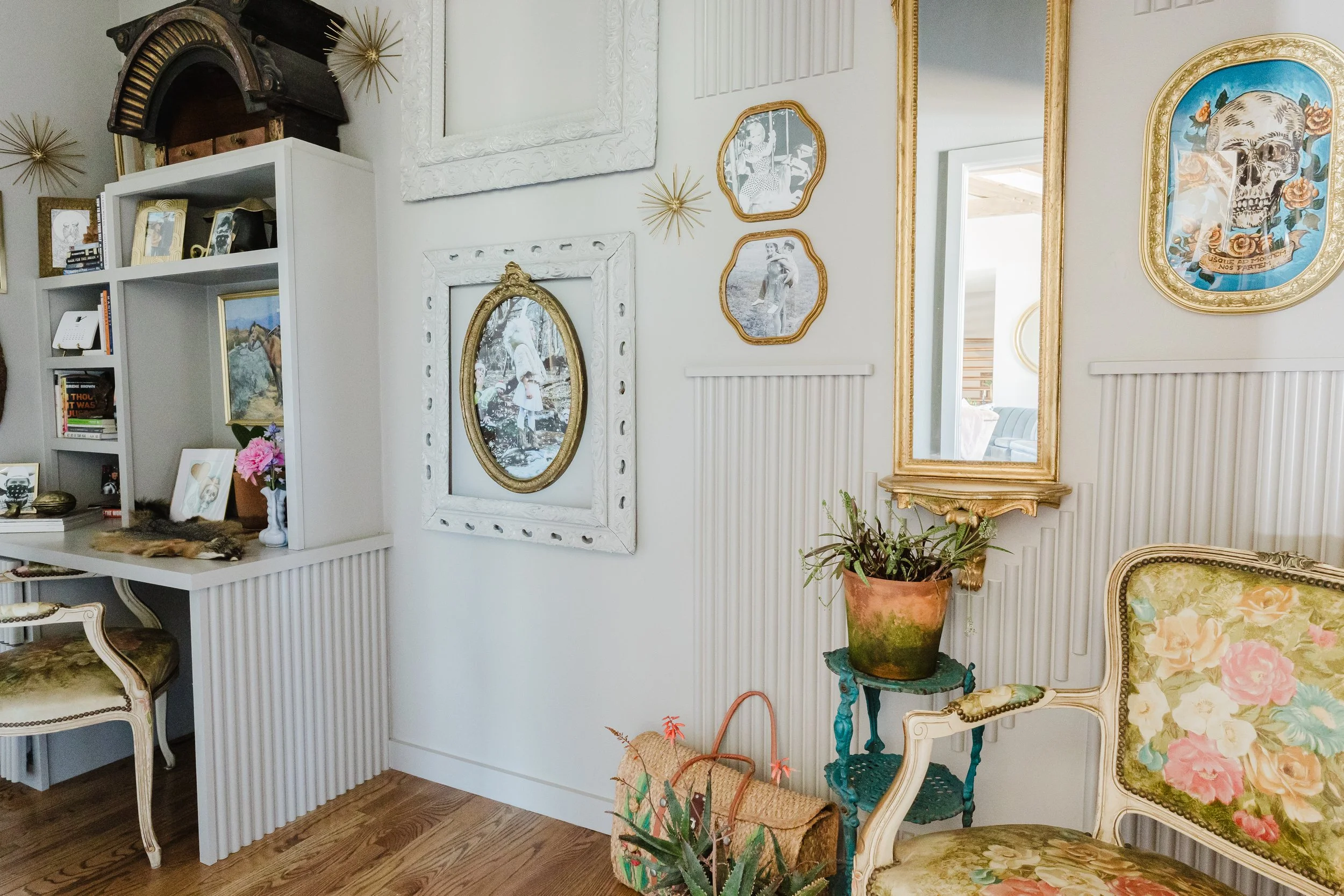 Living room wall decorated with various framed pictures, mirrors, and gold accents, including a skull artwork with floral elements, furniture with floral upholstery, a potted plant, and decorative items on shelves.