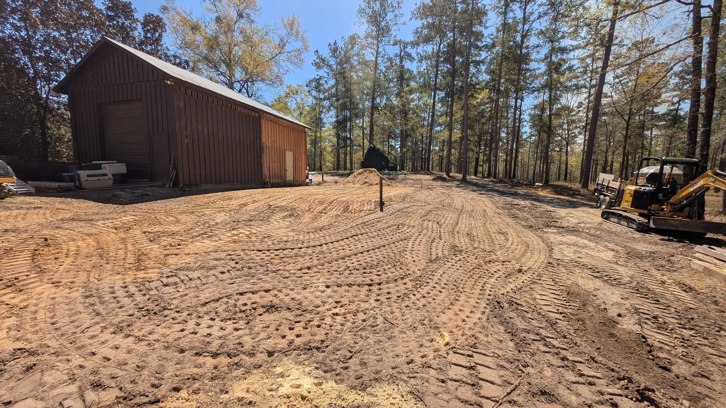 A construction site in a wooded area with a large wooden building, construction equipment, and dirt ground with tire tracks.