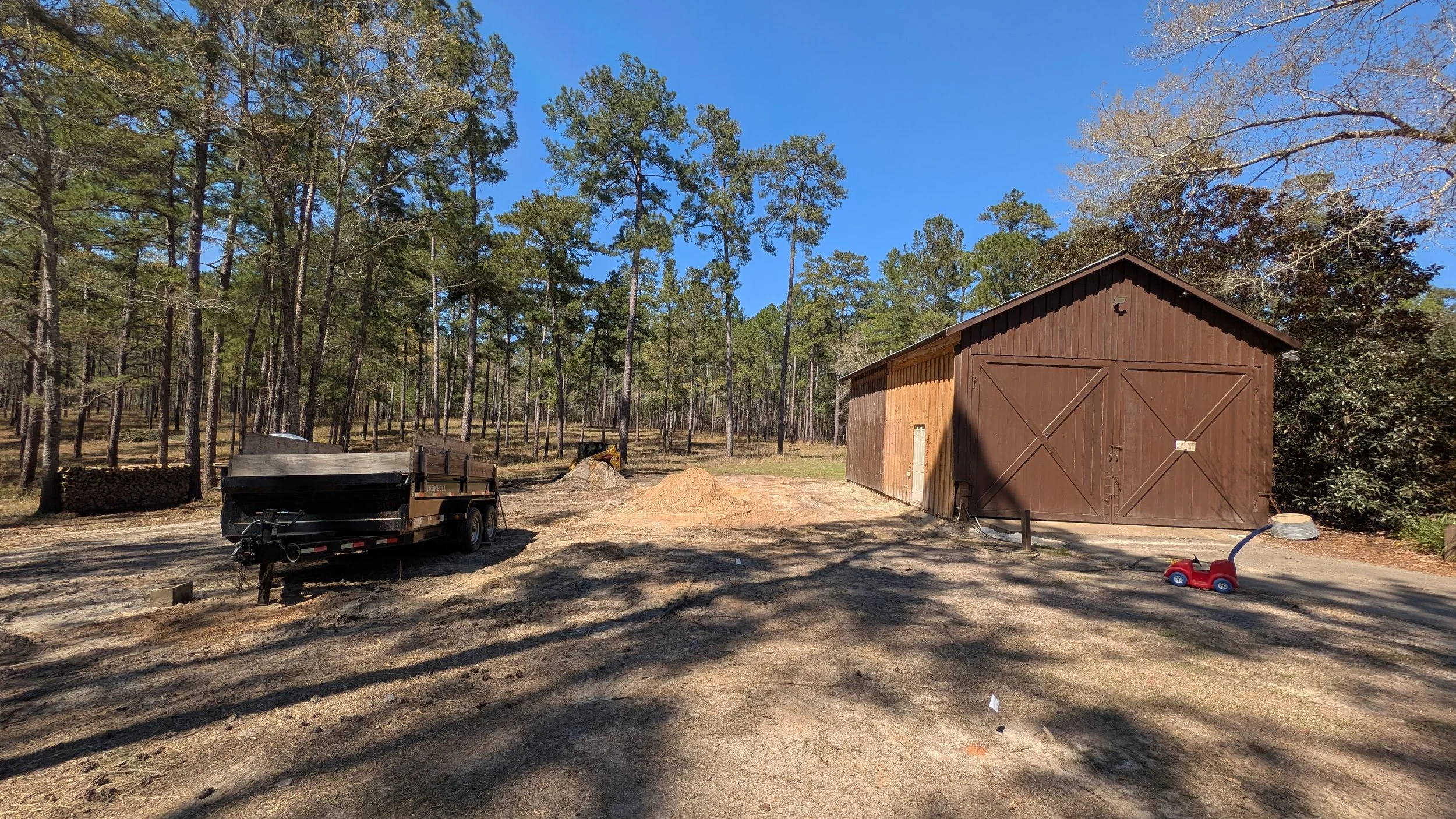A wooded outdoor area with a brown shed, a black trailer, and a red toy car. Piles of wood and dirt are nearby, with a partially cleared ground and tall pine trees in the background under a clear blue sky.