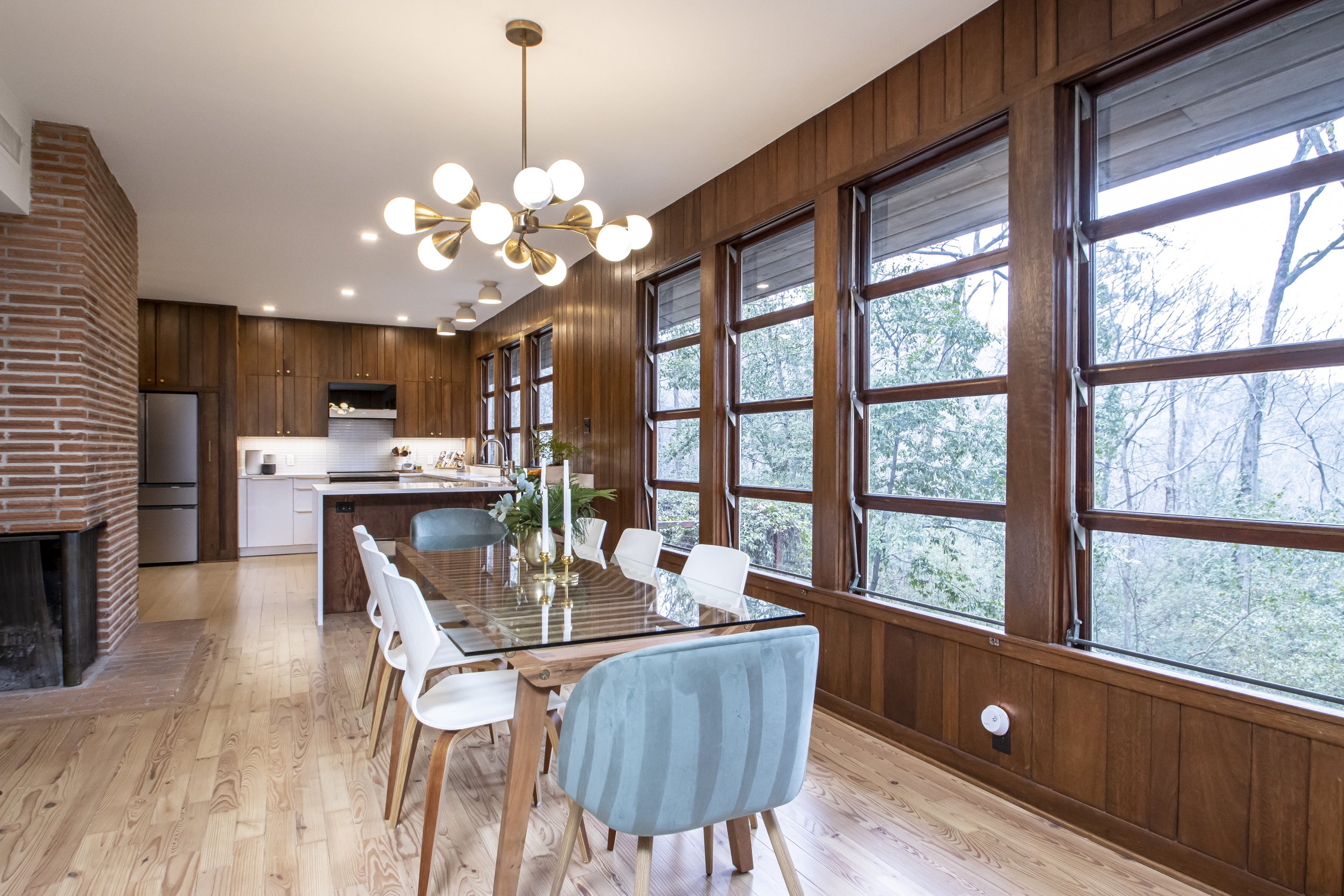A dining area with a glass-top table, white and light blue chairs, wood-paneled walls, large windows showing trees outside, a modern 12-bulb chandelier, and a kitchen with white cabinets and a brick fireplace on the side.