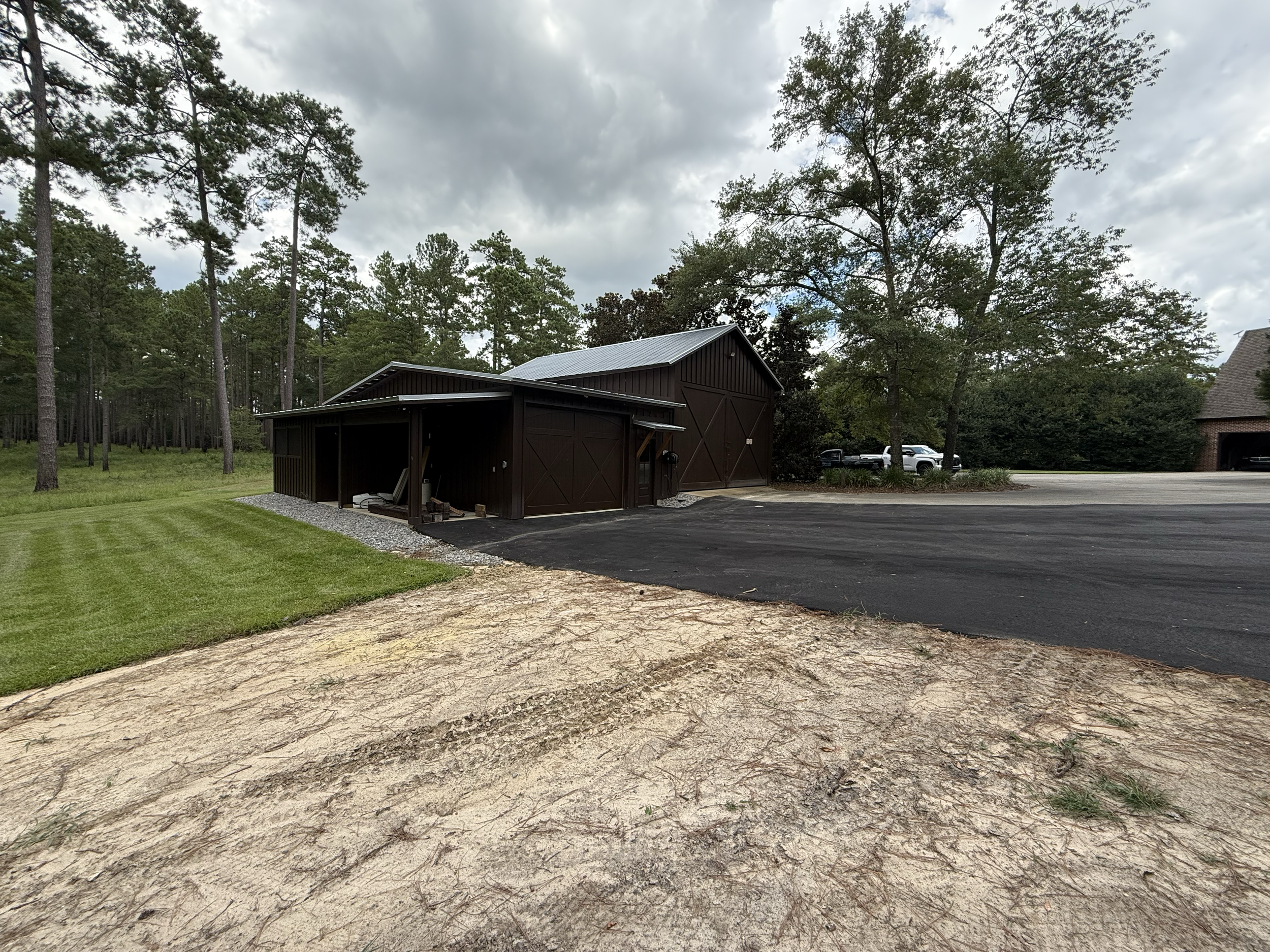 A rural scene with a brown metal barn surrounded by tall pine trees and some parked cars in the background, under a cloudy sky.