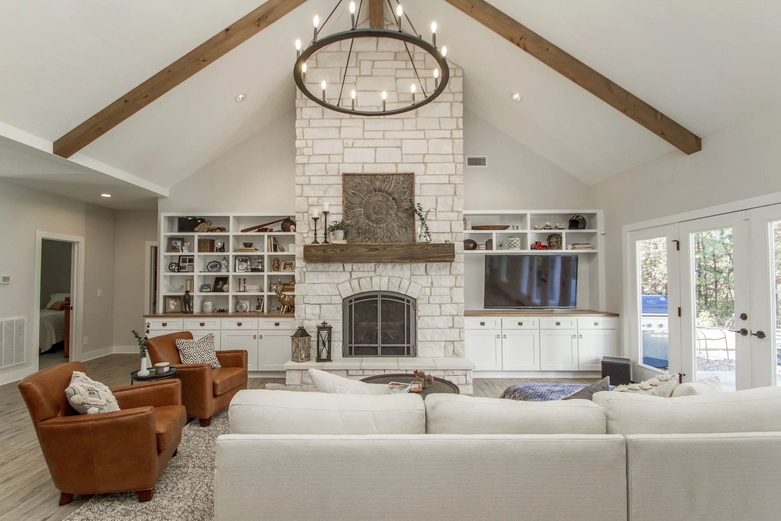 Living room with a high vaulted ceiling, a large brick fireplace with a wooden mantle, built-in white shelves on each side, a chandelier hanging from the ceiling, and furniture including a beige couch and two leather armchairs.