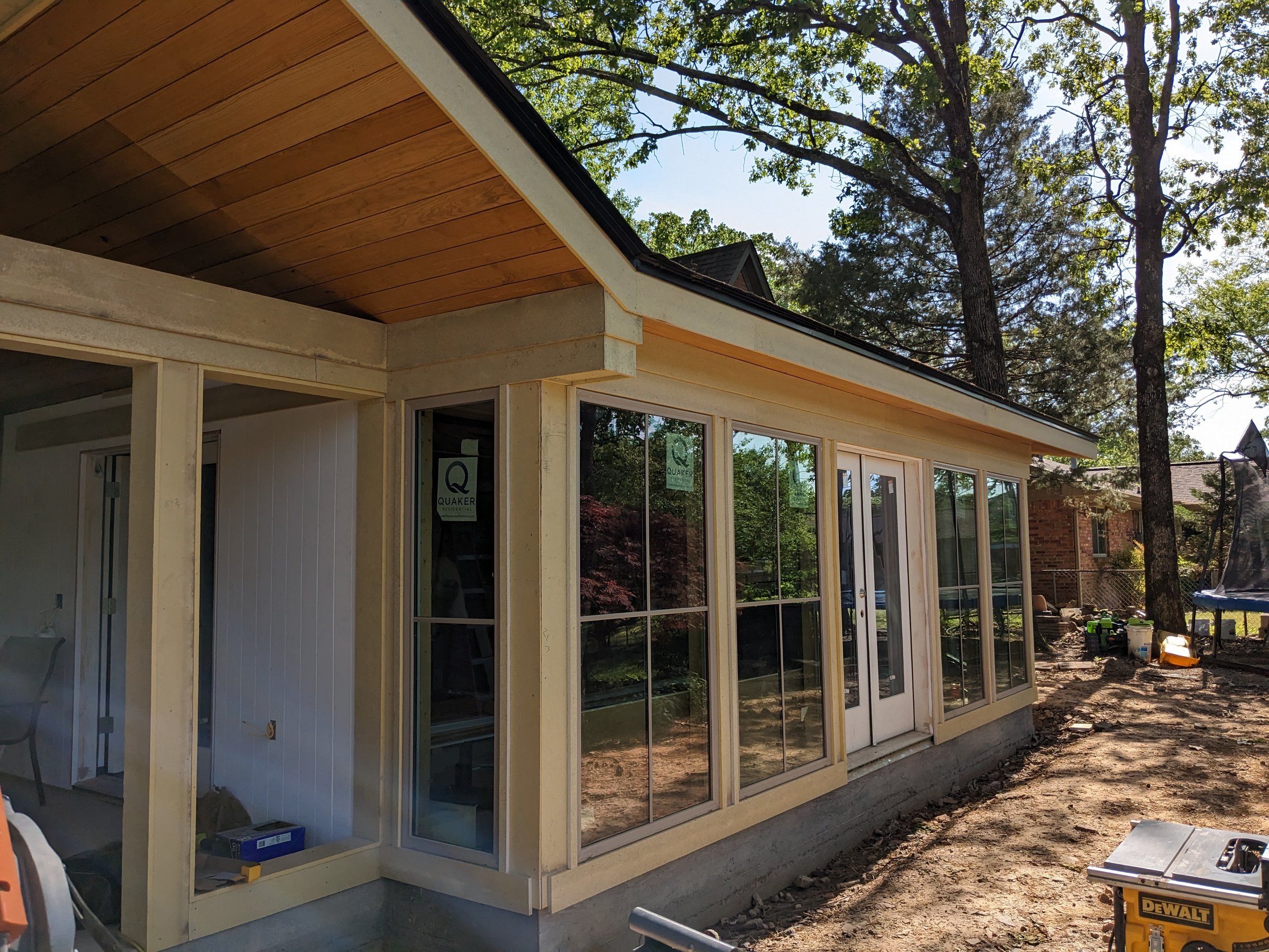 The exterior of a house under construction with large windows, a wooden roof overhang, and surrounded by trees.
