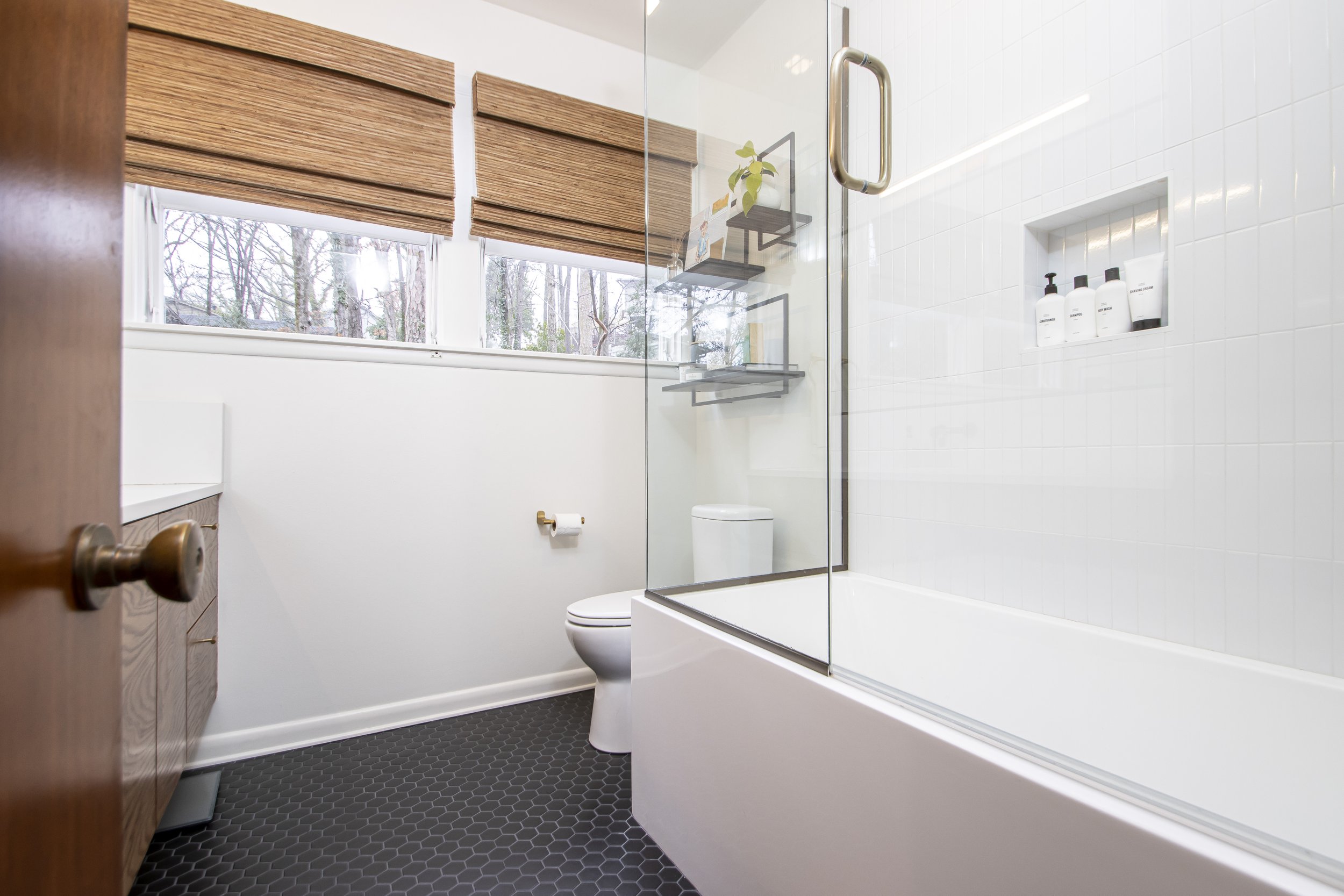 A modern bathroom featuring a white bathtub with a glass shower enclosure, a white toilet, dark hexagonal tile flooring, a wooden vanity with a brass handle, and windows with wooden blinds revealing leafless trees outside.