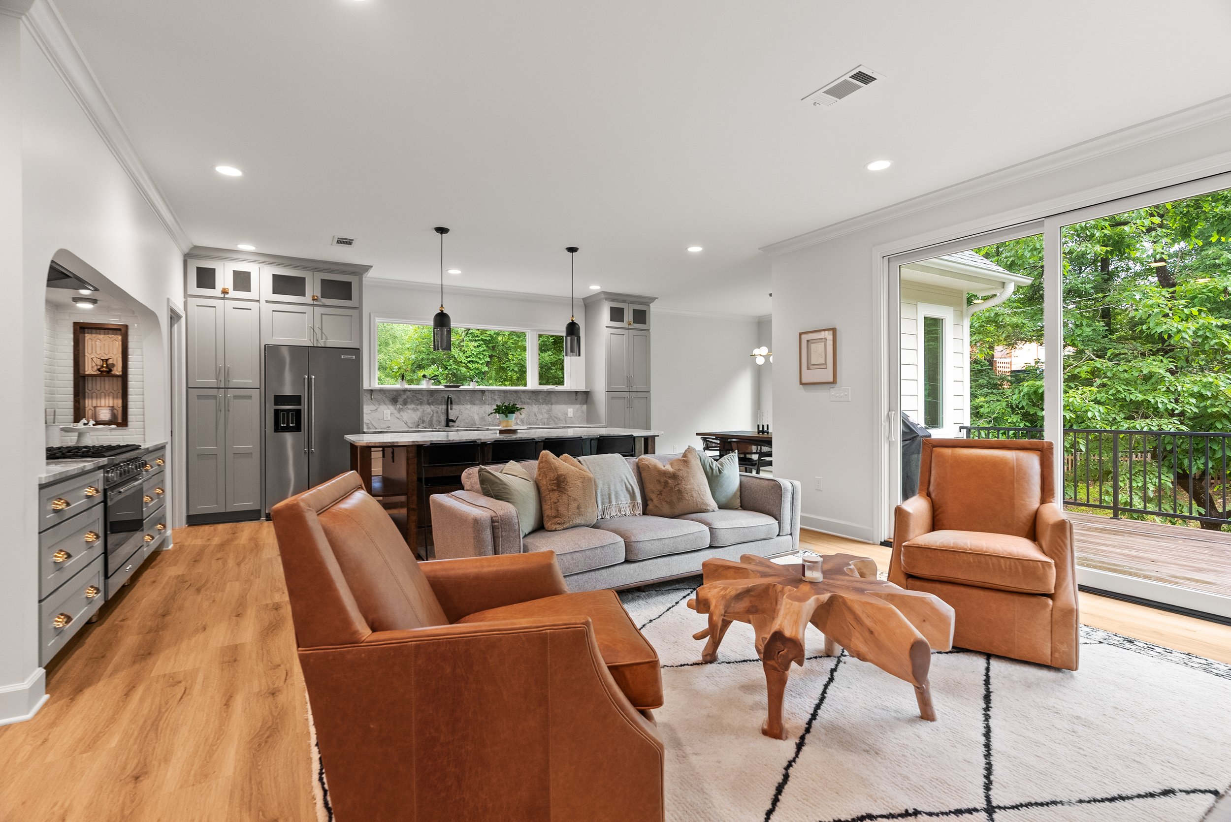 Open-concept living room and kitchen with large sliding glass door revealing a green outdoor area, featuring a gray sofa, leather armchairs, wooden coffee table, kitchen with gray cabinets, stainless steel refrigerator, and pendant lights.