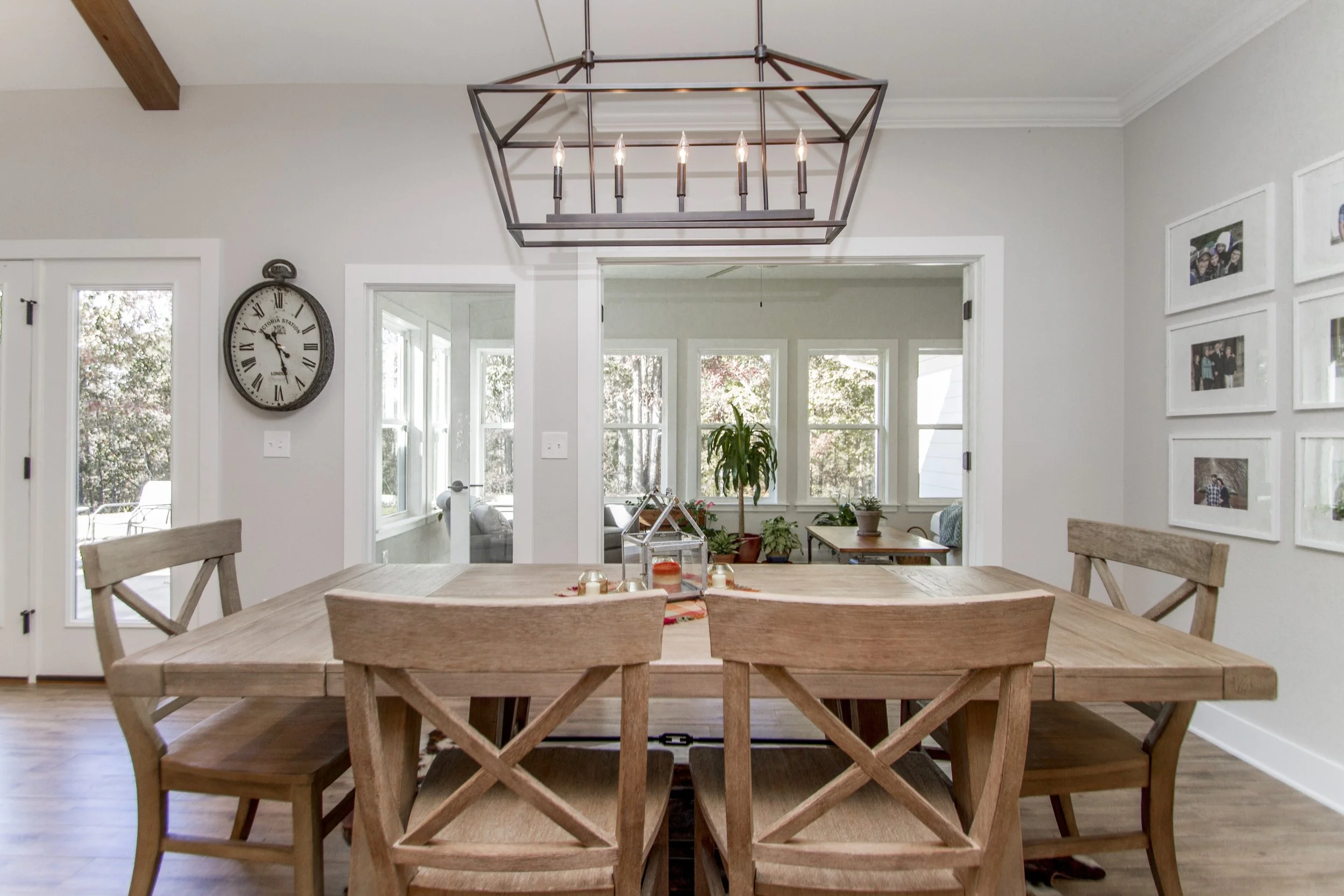 A dining room with a wooden table and chairs, a wall clock, a modern chandelier, and a background of windows and framed photographs.