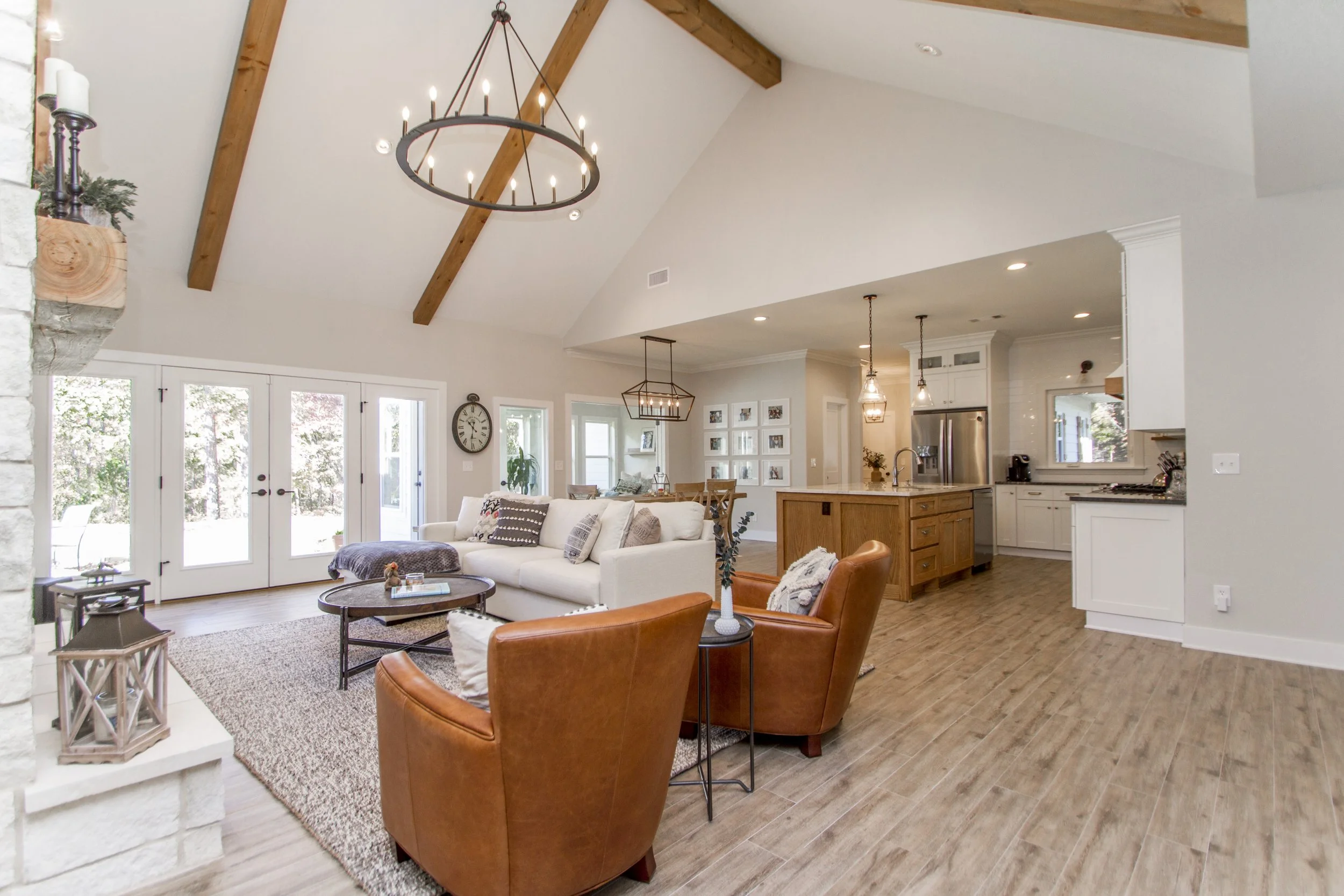 Living room with white sofa, leather armchairs, wooden ceiling beams, modern chandelier, coffee table, fireplace, large windows, and an open kitchen area.