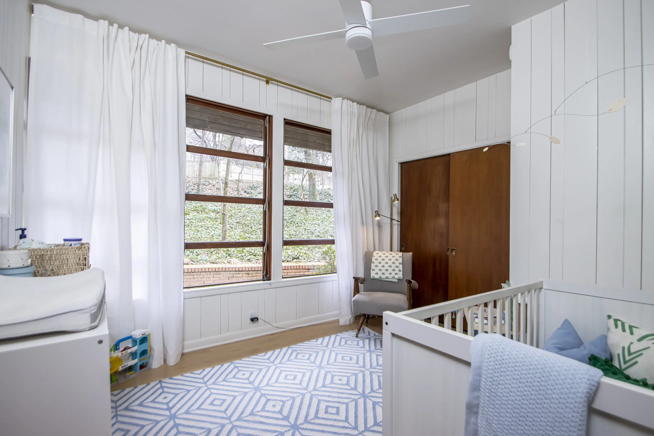 A nursery with white paneling, large windows with white curtains, a gray armchair near the window, and a crib in the foreground. There is a white and blue geometric patterned rug on the wooden floor.