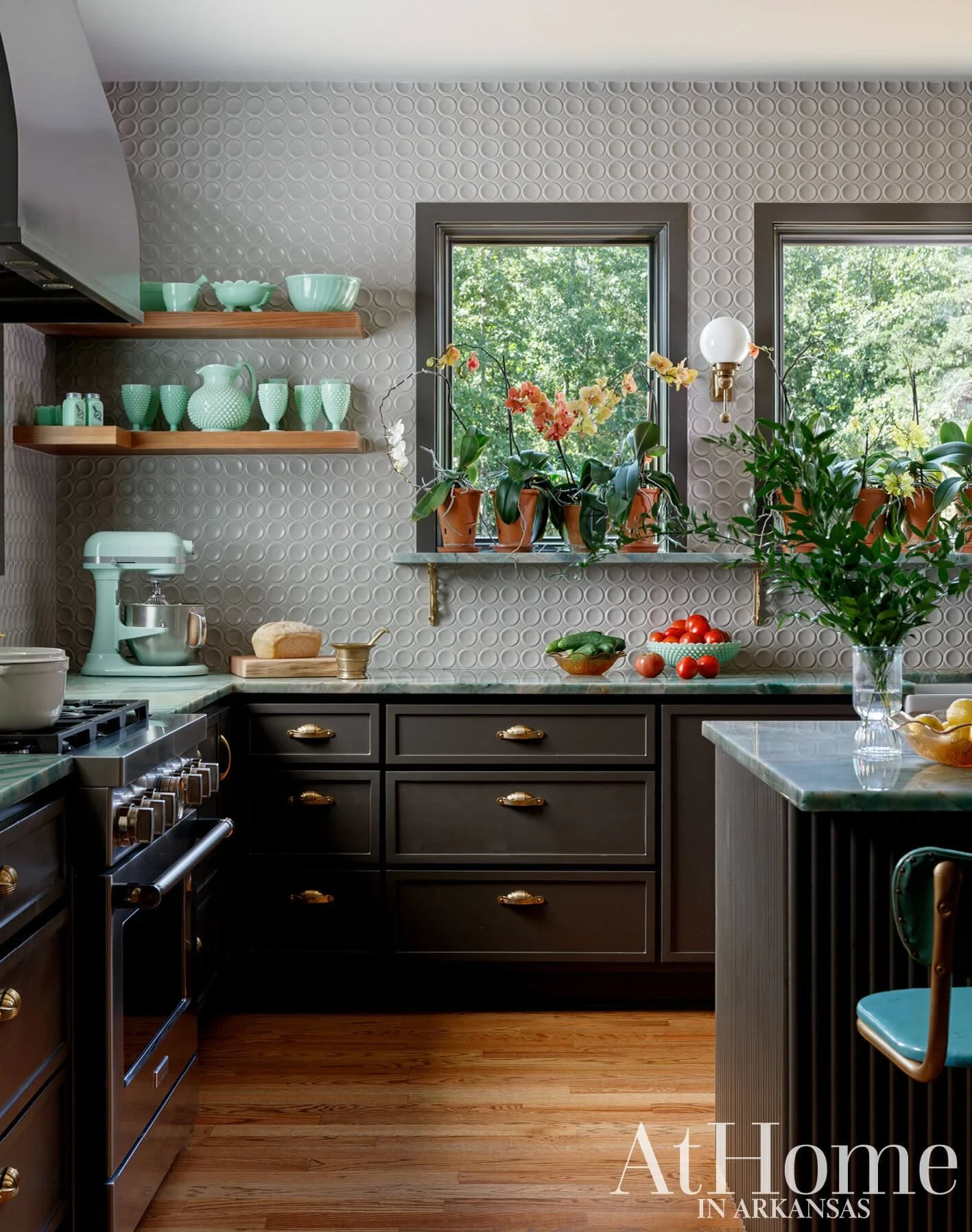 Kitchen with dark gray cabinets, green and white dishware on wooden shelves, orchids and other plants on the windowsill, a marble countertop with tomatoes and cucumbers, and a window showing green trees outside.