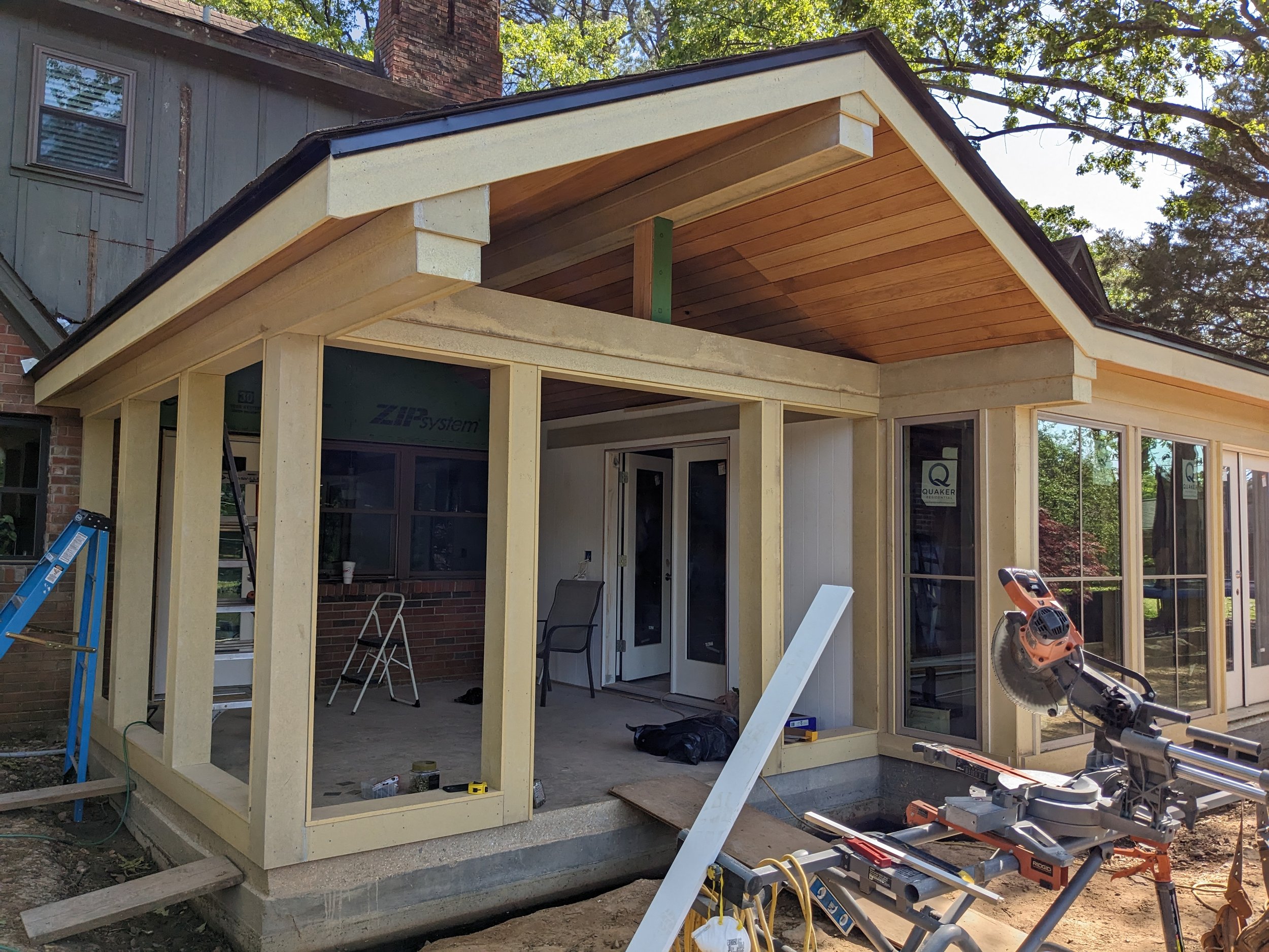 Construction site where a new porch or extension is being built onto a house. The framing is in place with some wooden ceiling panels installed. Construction tools and materials are scattered around the area.