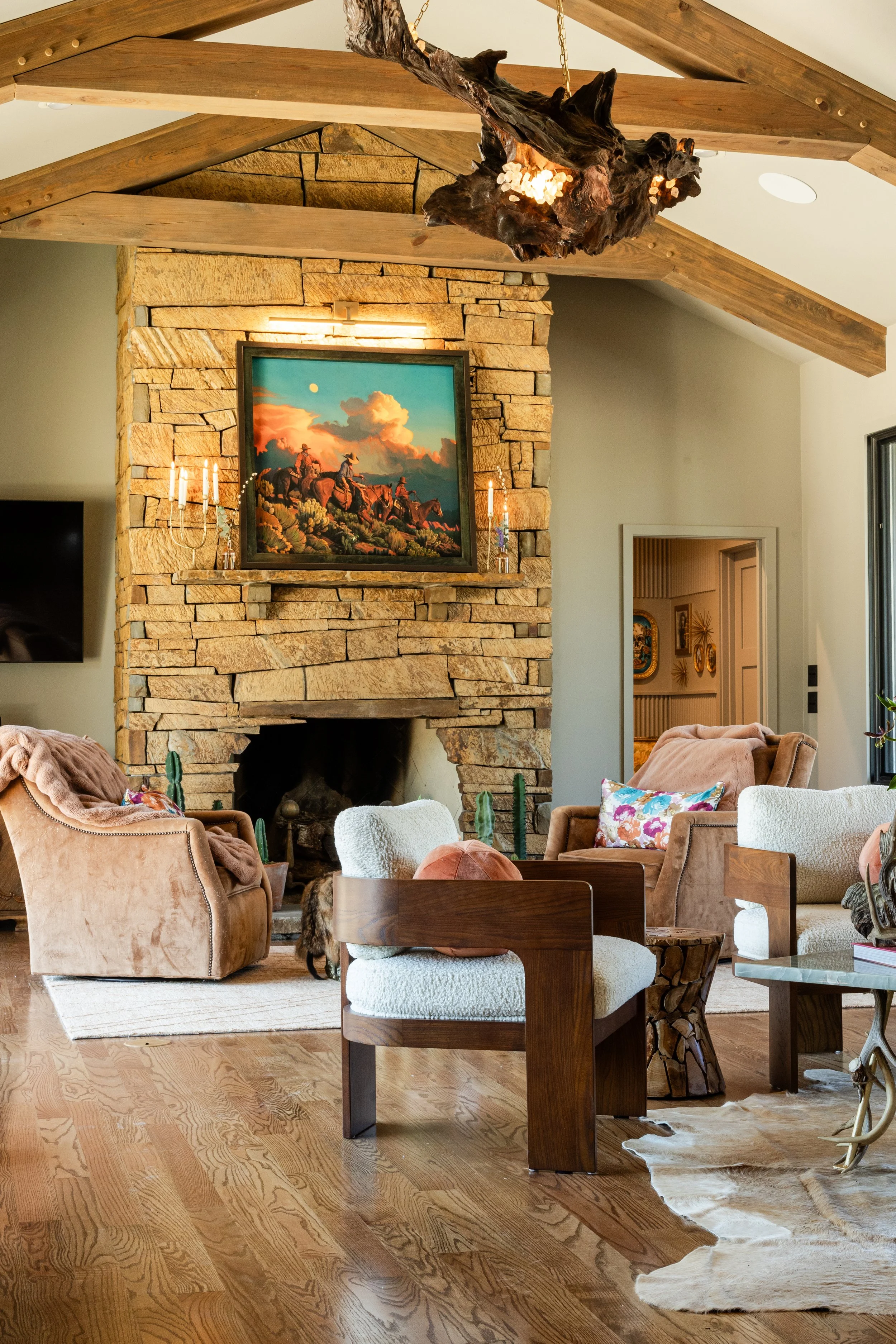 Living room with a stone fireplace, a large landscape painting of cowboys on horseback under a cloudy sky, various armchairs and sofas, a cowhide rug, and a wooden table and chairs, all with warm lighting and wooden ceiling beams.