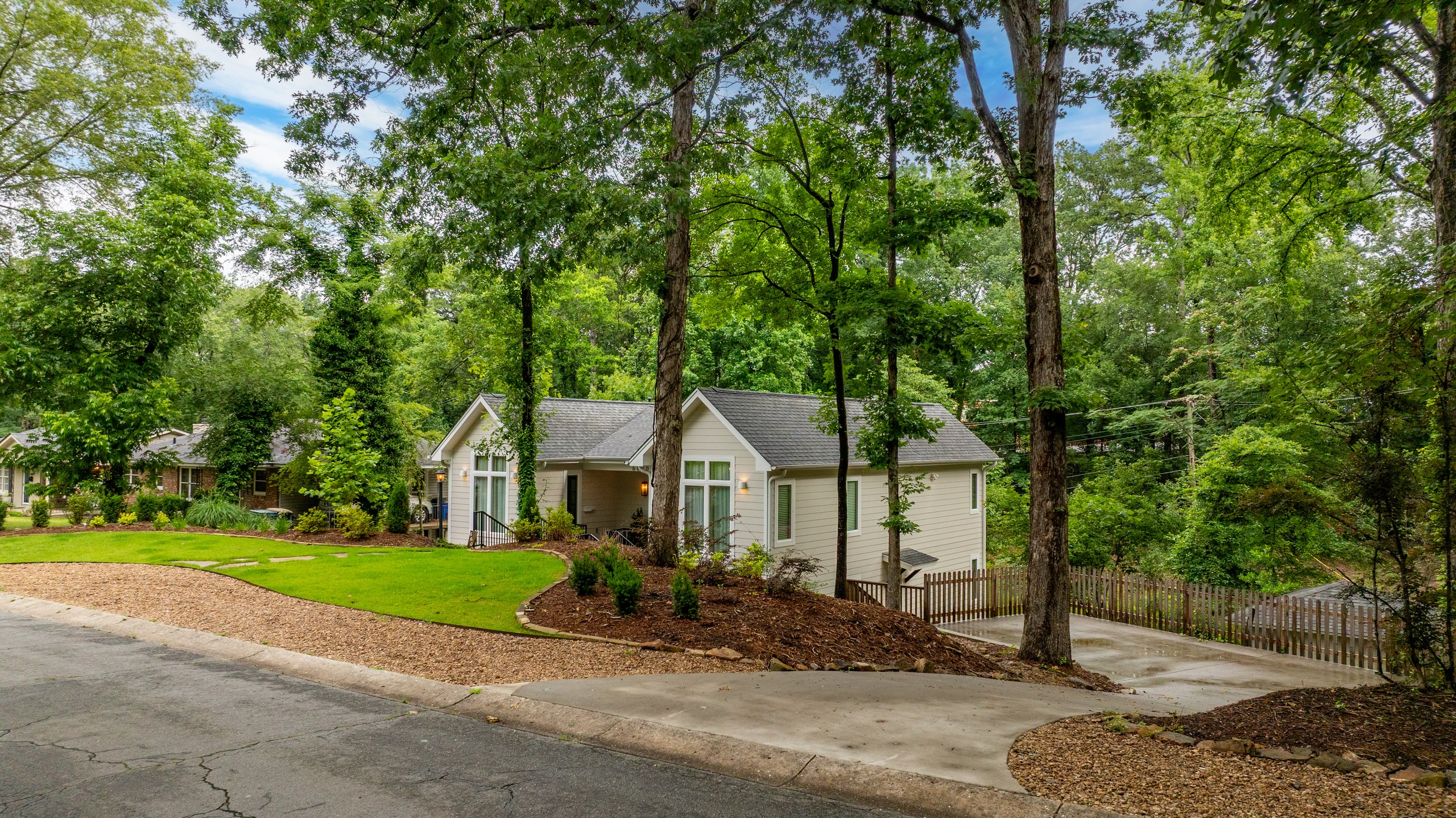 Suburban neighborhood scene with a white house amidst tall trees, a neatly maintained lawn, and a curved driveway, under a partly cloudy sky.