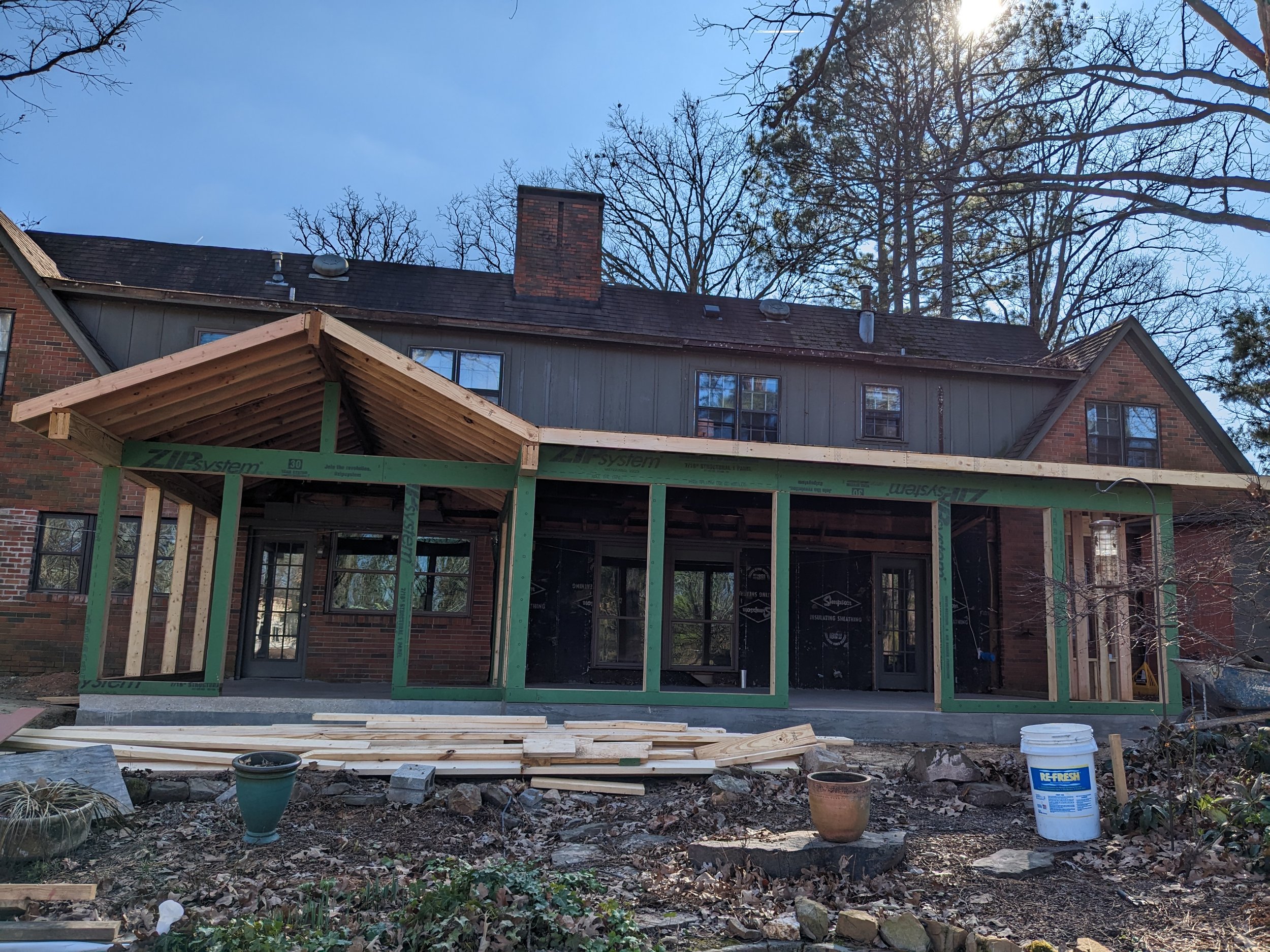 A residential construction site showing the back of a house with a new wooden roof structure being built, and the house has brick and siding exterior. There are construction materials and buckets on the ground, and leafless trees with the sun shining