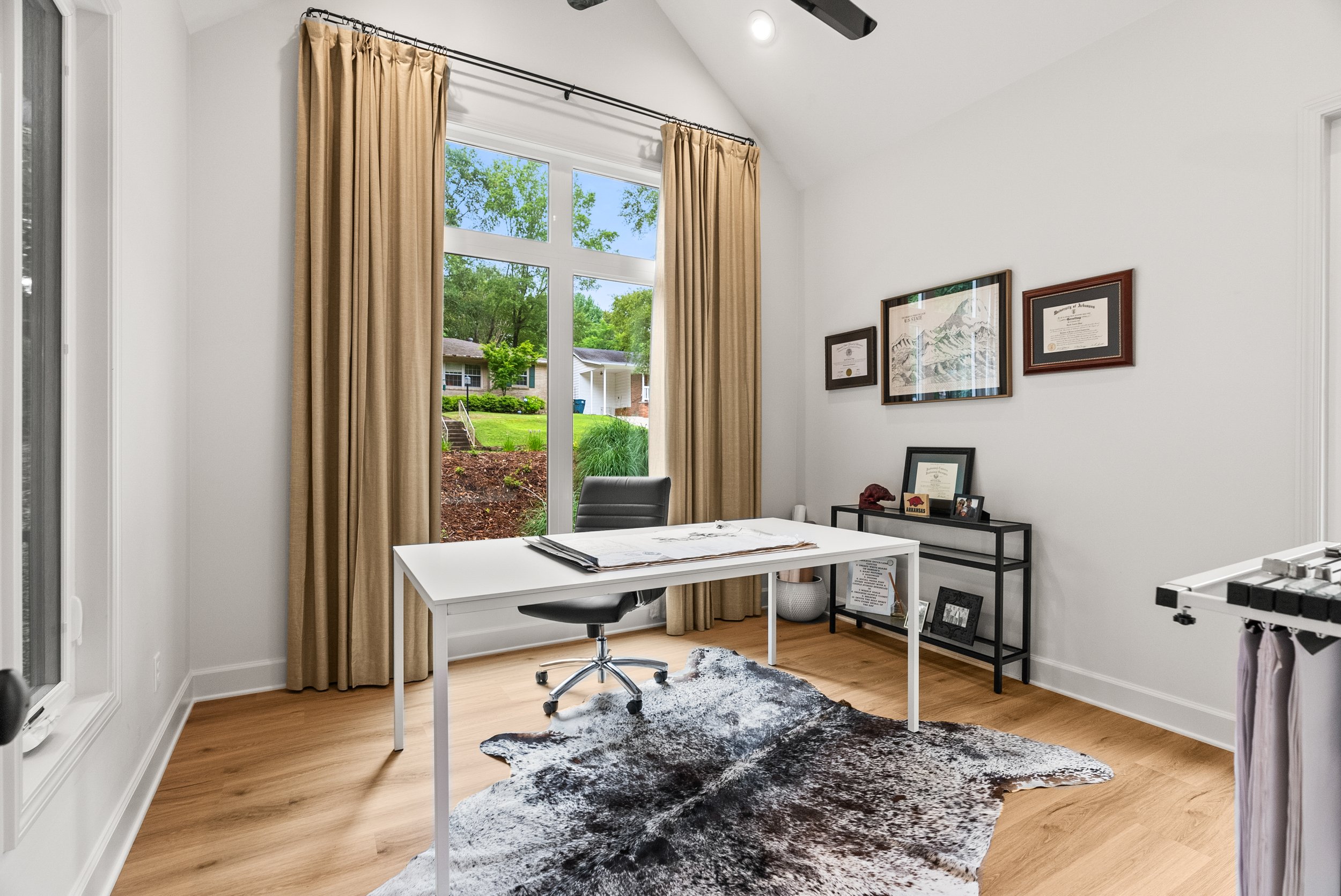 Home office with large window, beige curtains, white desk with papers, black office chair, black framed diplomas on wall, black shelving with framed photos, cowhide rug, and foosball table