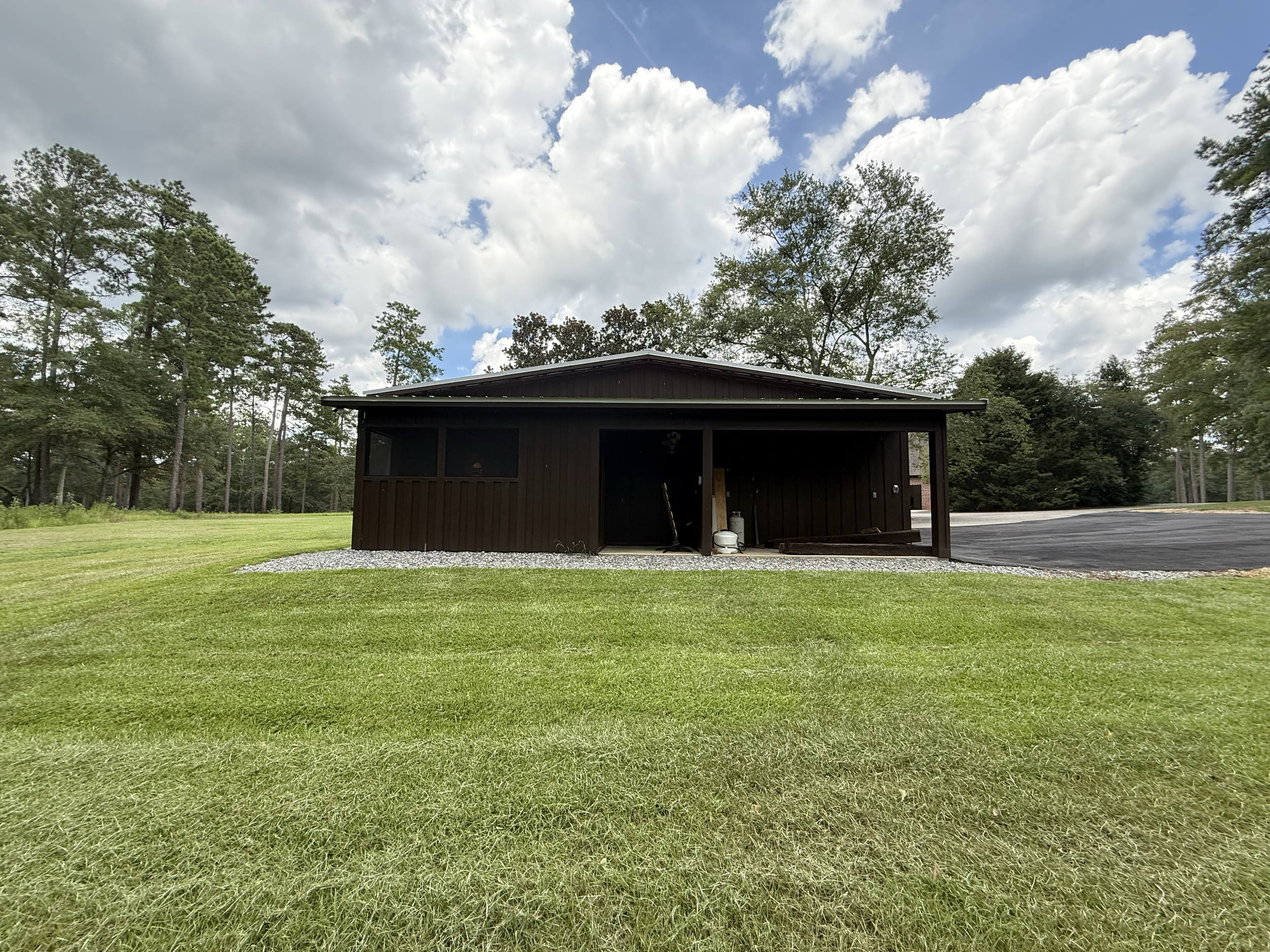 A black wooden building with an open front, surrounded by a grassy lawn and tall trees, under a partly cloudy sky.