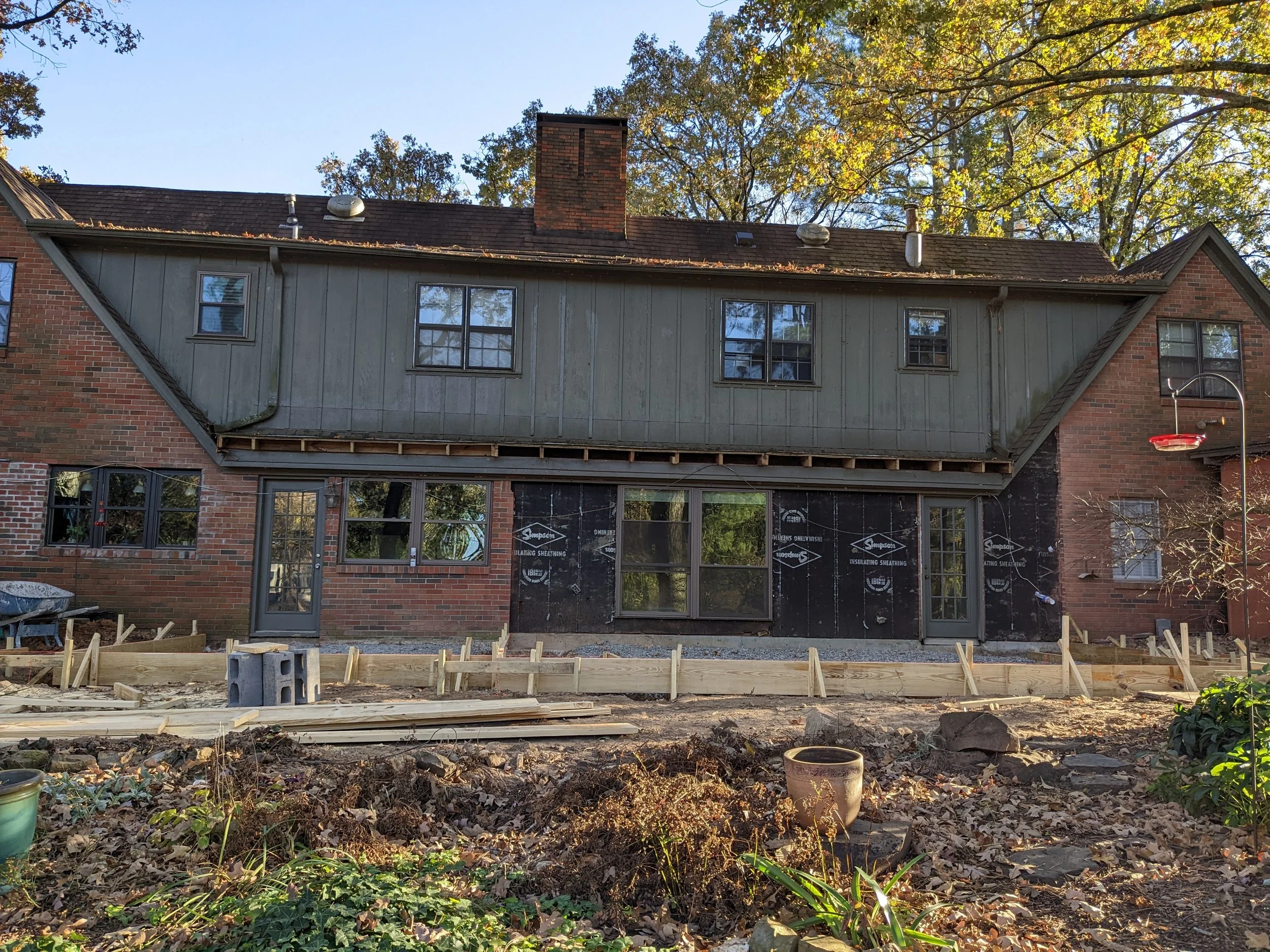 Back view of a house under construction with wooden framing and brick walls, surrounded by trees with yellow blooms and a clear blue sky.