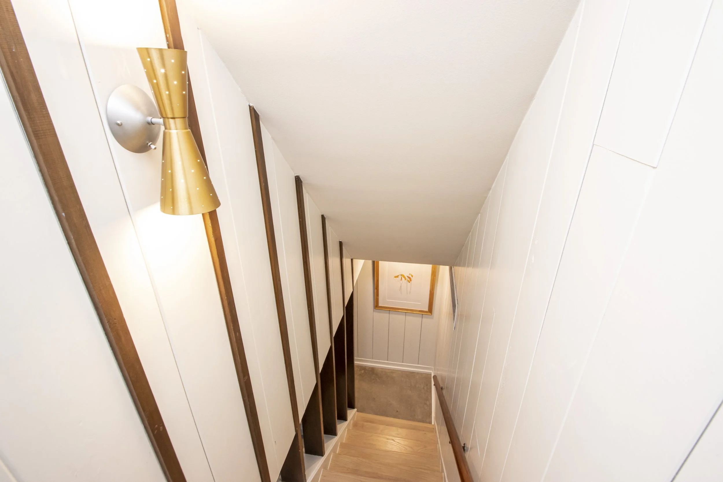 Interior view of a staircase with wooden treads, white paneled walls, and a handrail on the right side. The ceiling slopes downward, and there is a framed picture or mirror on the wall at the bottom of the stairs.