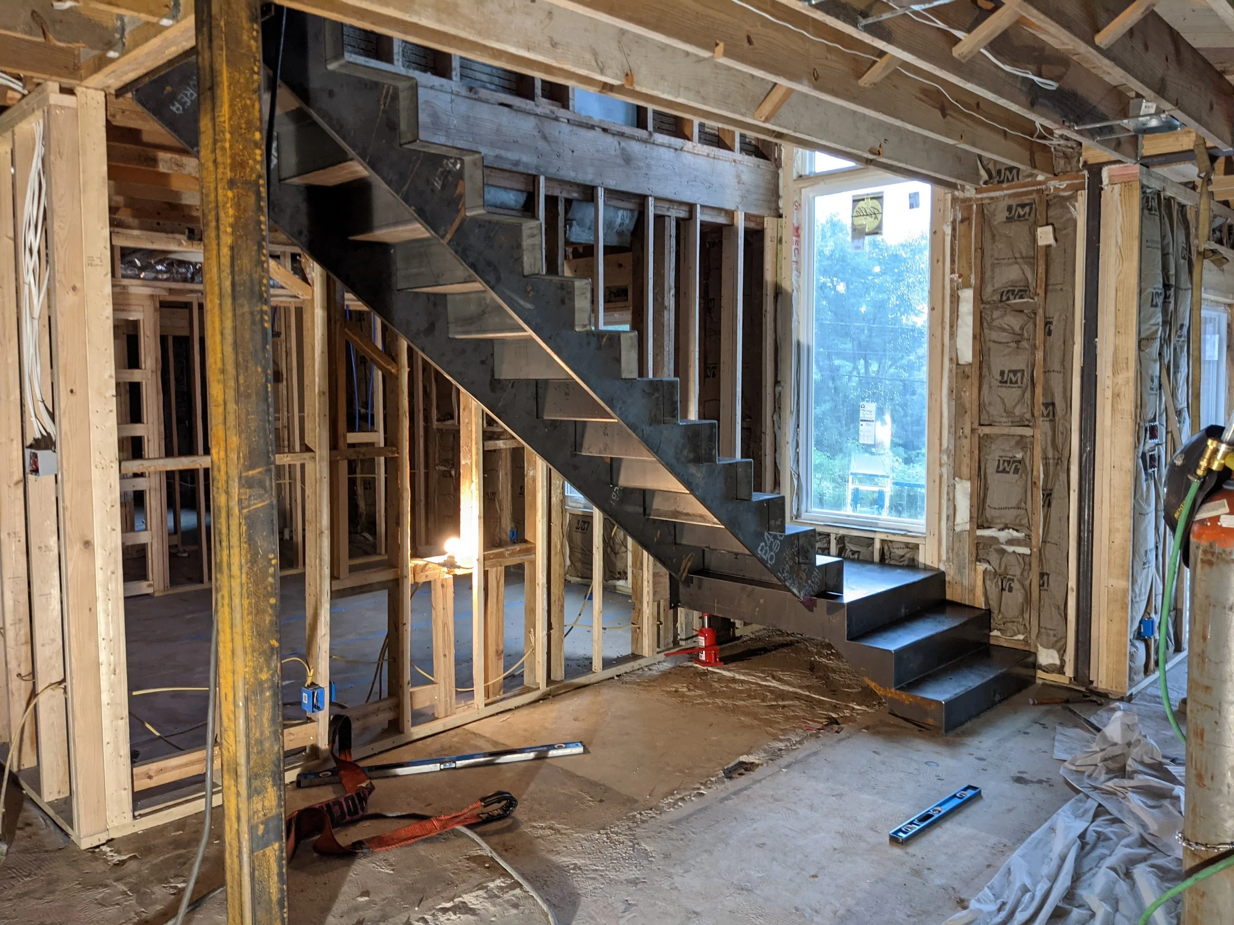 Interior of a house under construction with exposed wooden framing, staircase, and large window.
