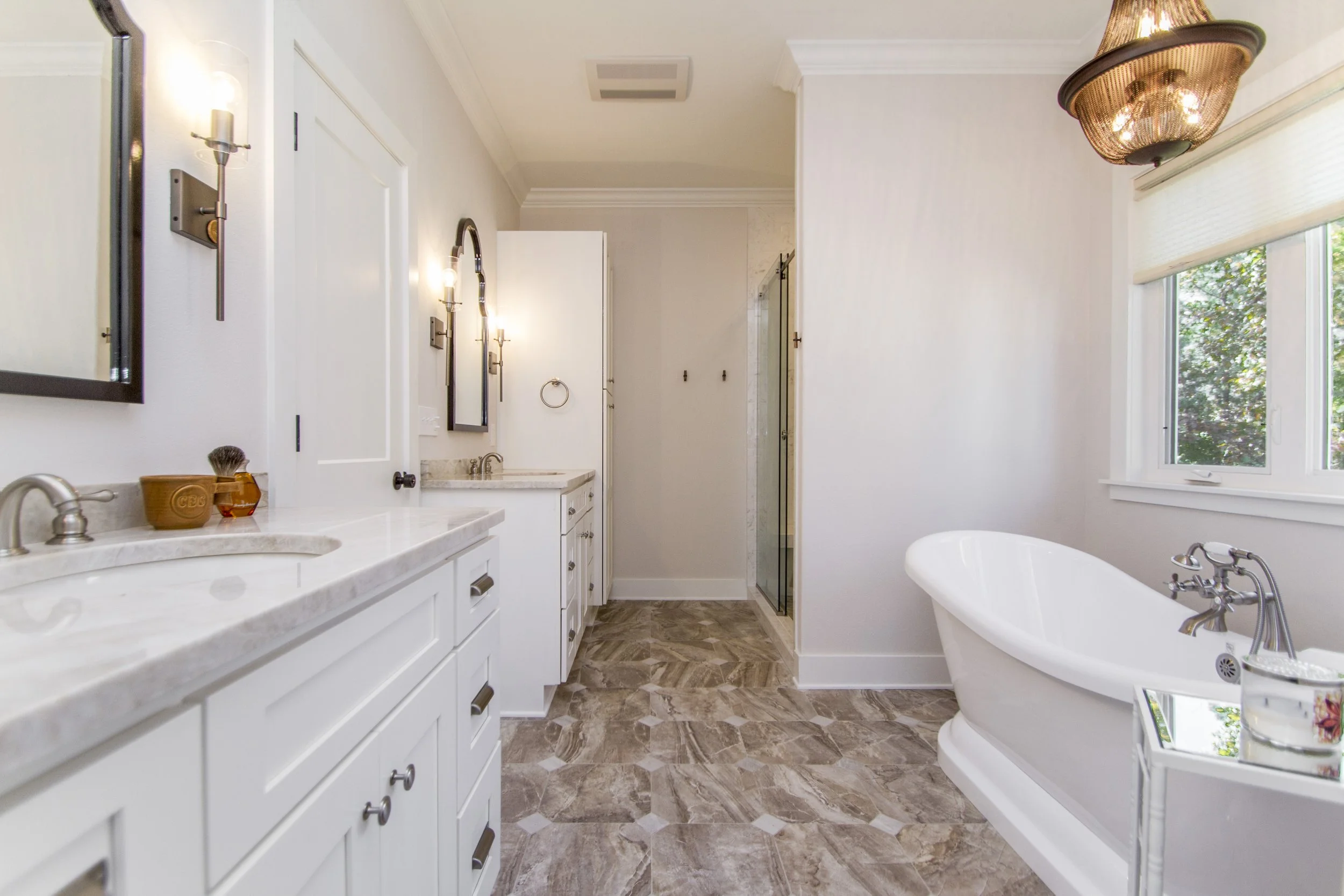 A spacious bathroom with white cabinets, a large bathtub, and a shower with glass door. The bathroom has a window with blinds, decorative wall fixtures, and a marble countertop.