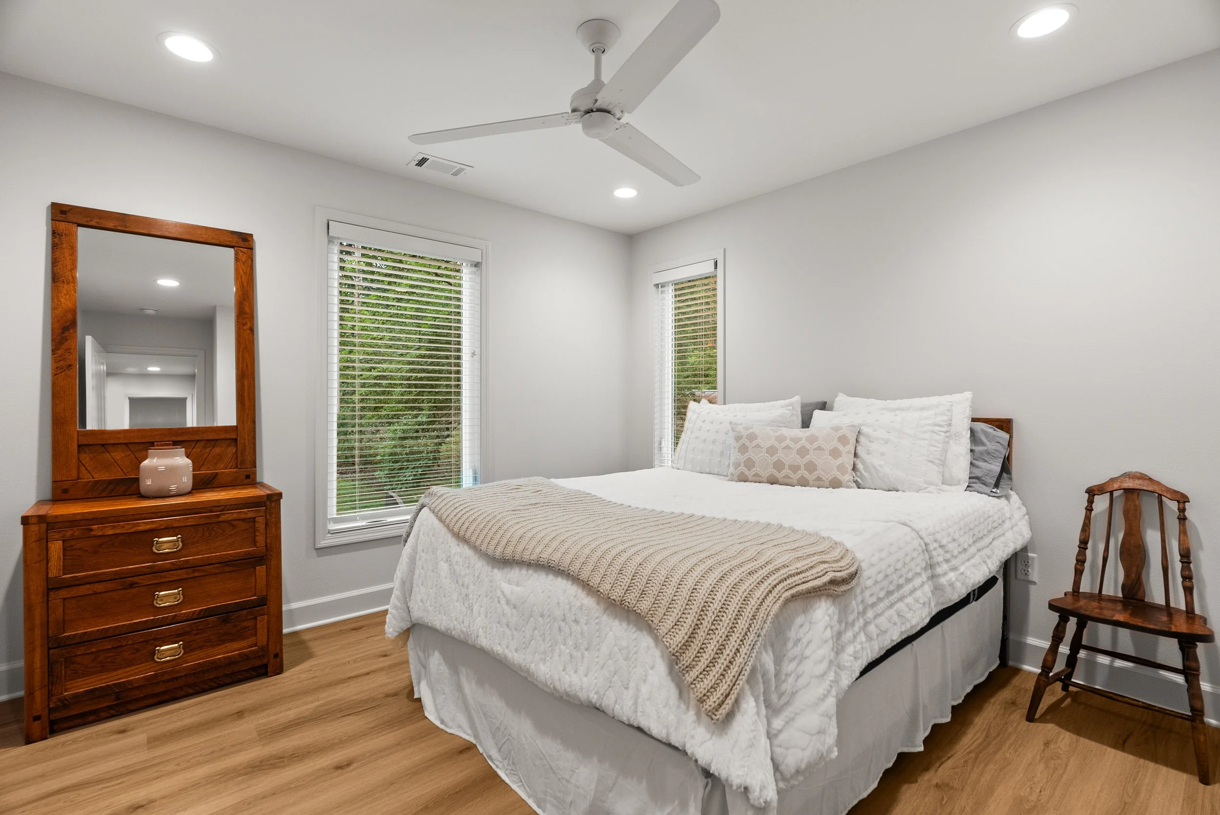 The image shows a cozy bedroom with a white bed dressed in a textured white comforter and pillows. A beige knit blanket is draped over the foot of the bed. To the left, there is a wooden dresser with a mirror and a pink vase on top. Two windows with 
