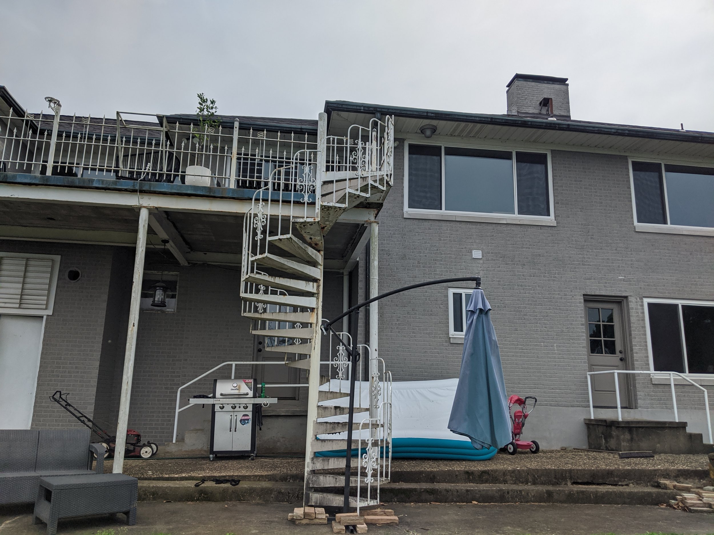 Backyard patio with outdoor stairs leading to a second-story balcony, grill, pool, and various outdoor items in front of a gray brick house with large windows.